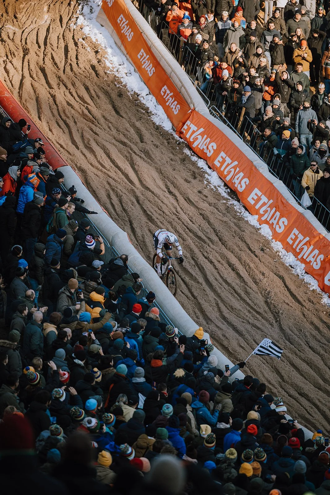 Cyclist riding on a dirt track during a race with a large crowd of spectators watching from the sidelines, some taking photos, and protective barriers separating them from the track. Snow is visible on the ground along the edges.