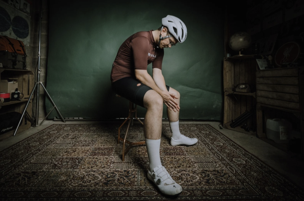 A man sitting on a stool in front of a green screen, wearing cycling gear including a helmet, glasses, cycling jersey, shorts, socks, and shoes, with a room containing various equipment and shelves in the background.