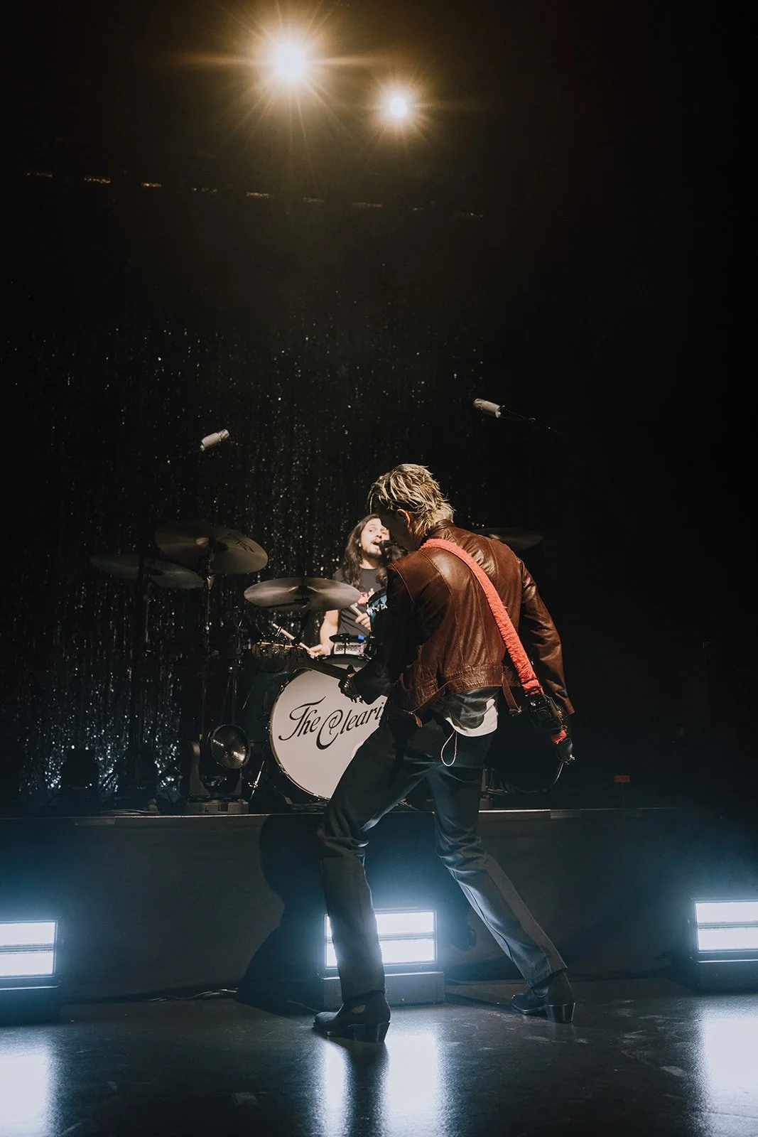 A live music performance featuring a guitarist in the foreground and a drummer in the background, with stage lights illuminating the scene.