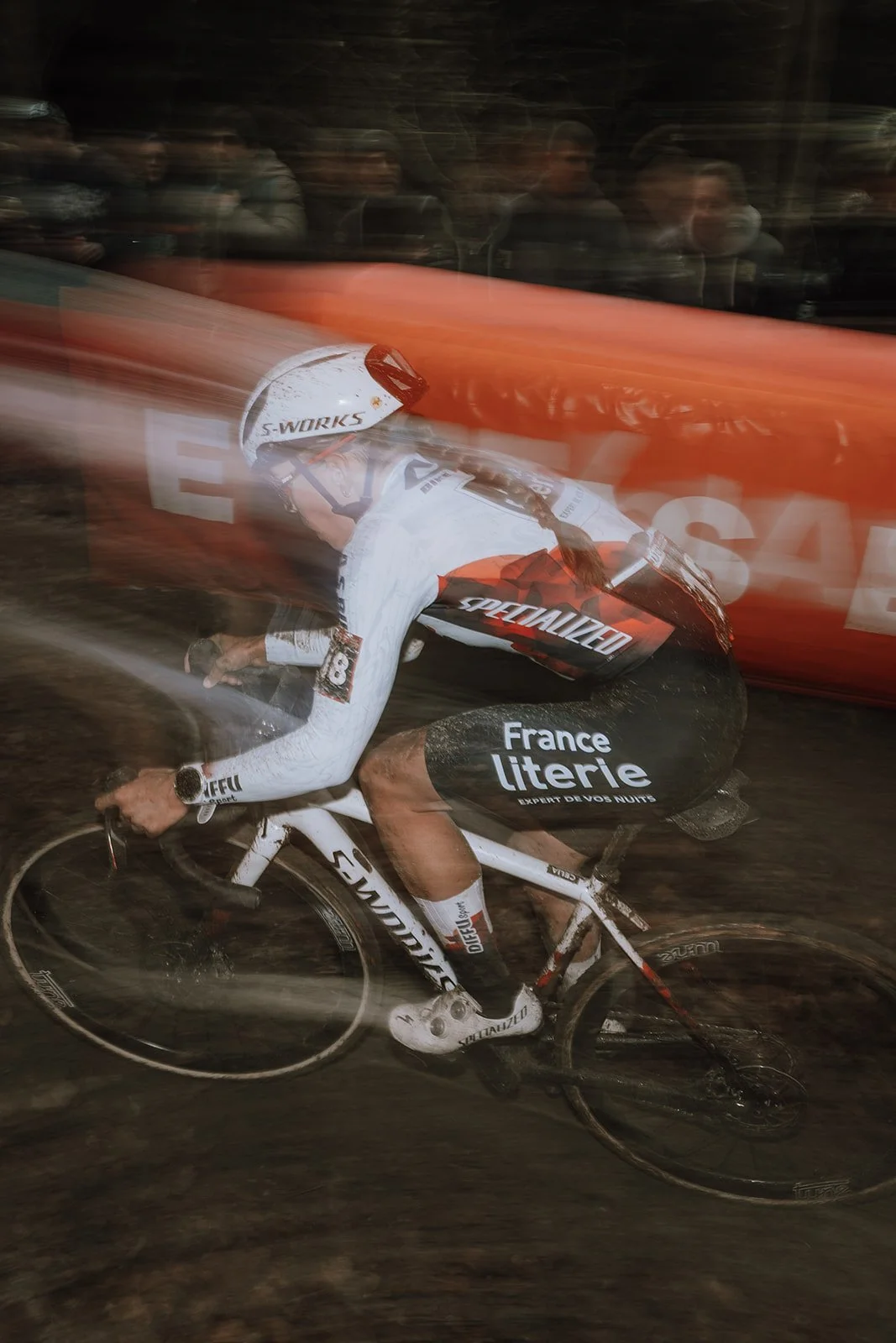A cyclist wearing a white and black racing uniform with logo branding, riding a specialized bike during a race at night with spectators blurred in the background.