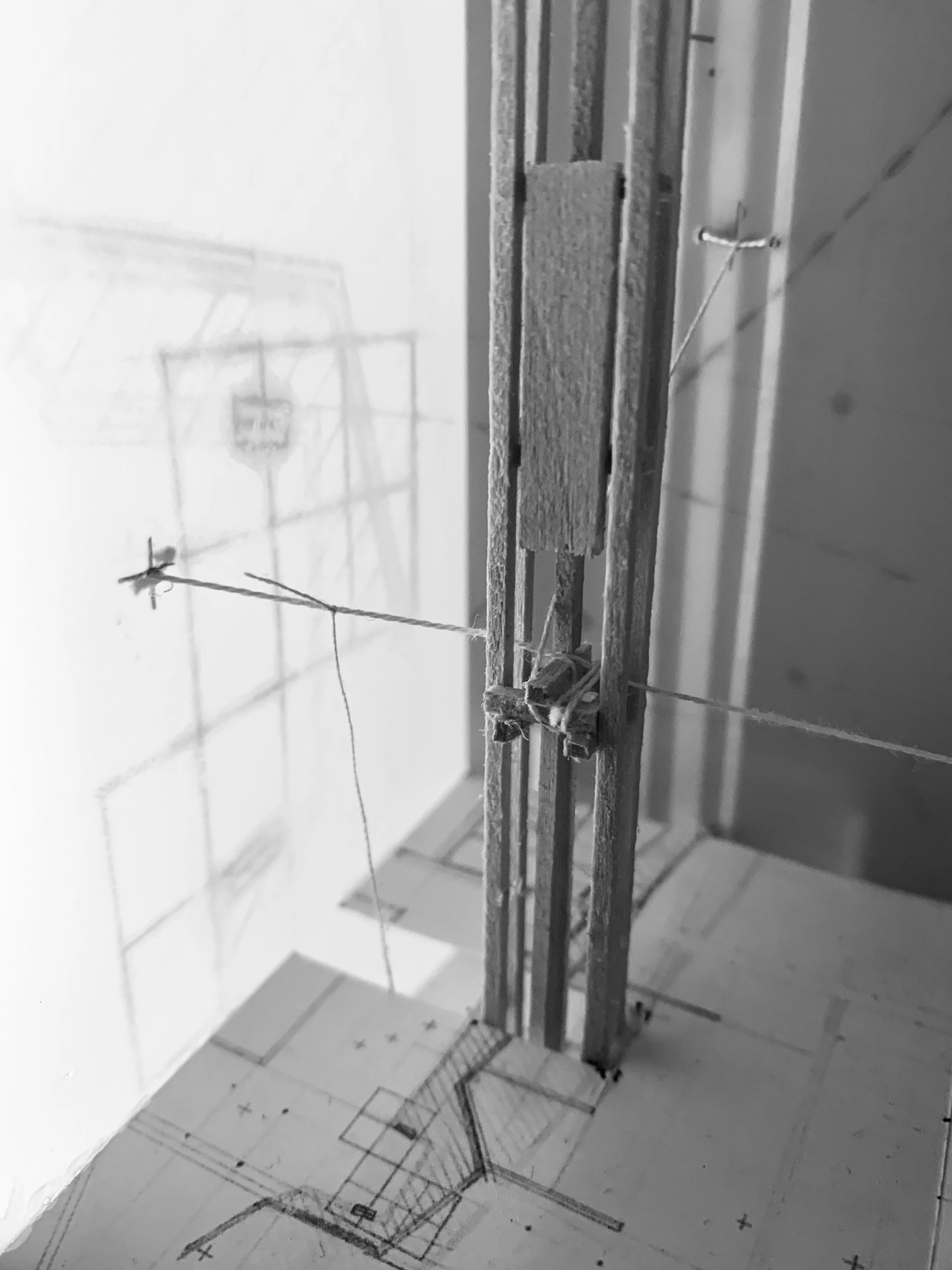 Construction site with a detailed blueprint on the floor, a tall structure with metal and wooden elements, and wires, with a shadow of a crane or lifting device cast on the wall.