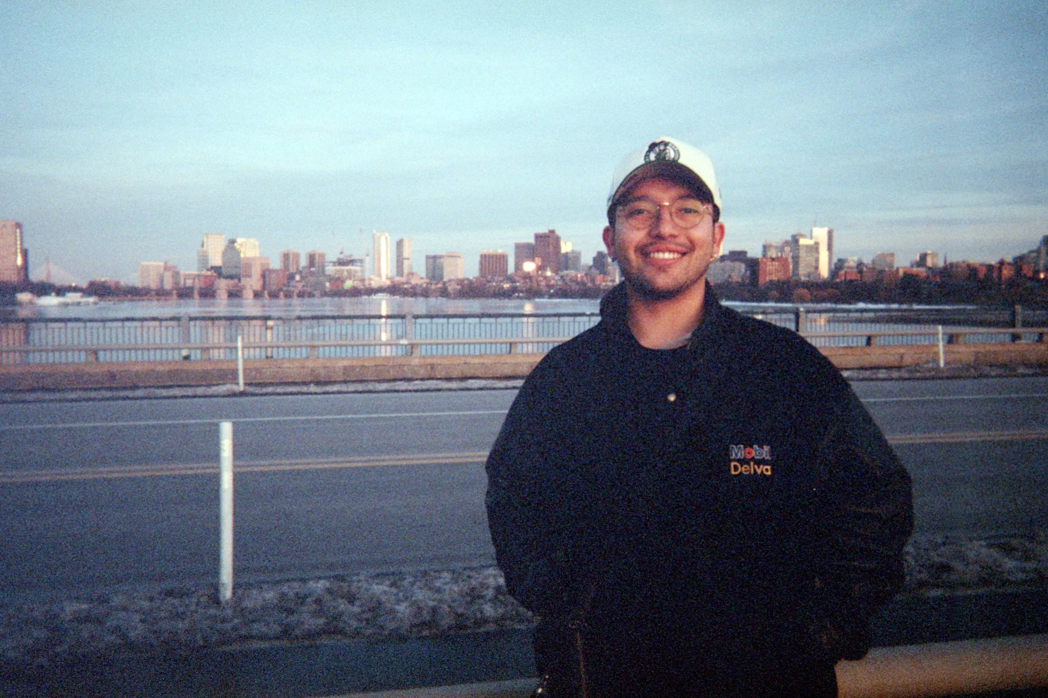 A smiling man wearing a black jacket with 'Mobil Delva' logo and a white cap, standing outdoors near a railing with a city skyline and river in the background.
