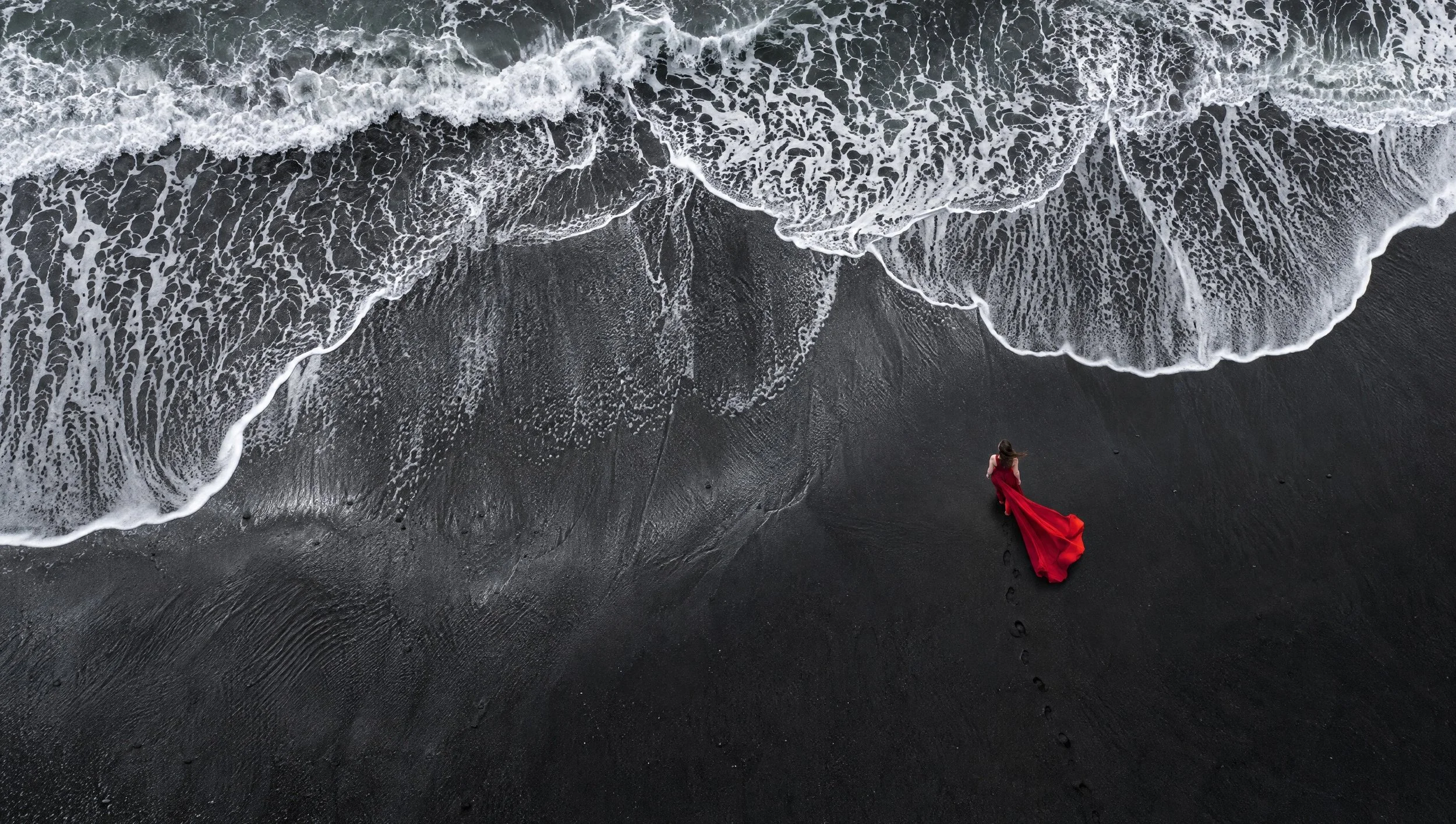 Aerial view of a woman in a crimson dress on a black sand beach.