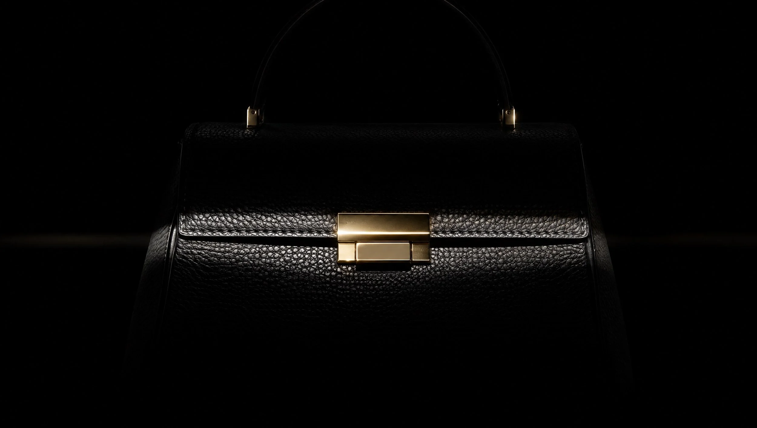A black leather handbag with a gold clasp, lit by a thin blade of light.