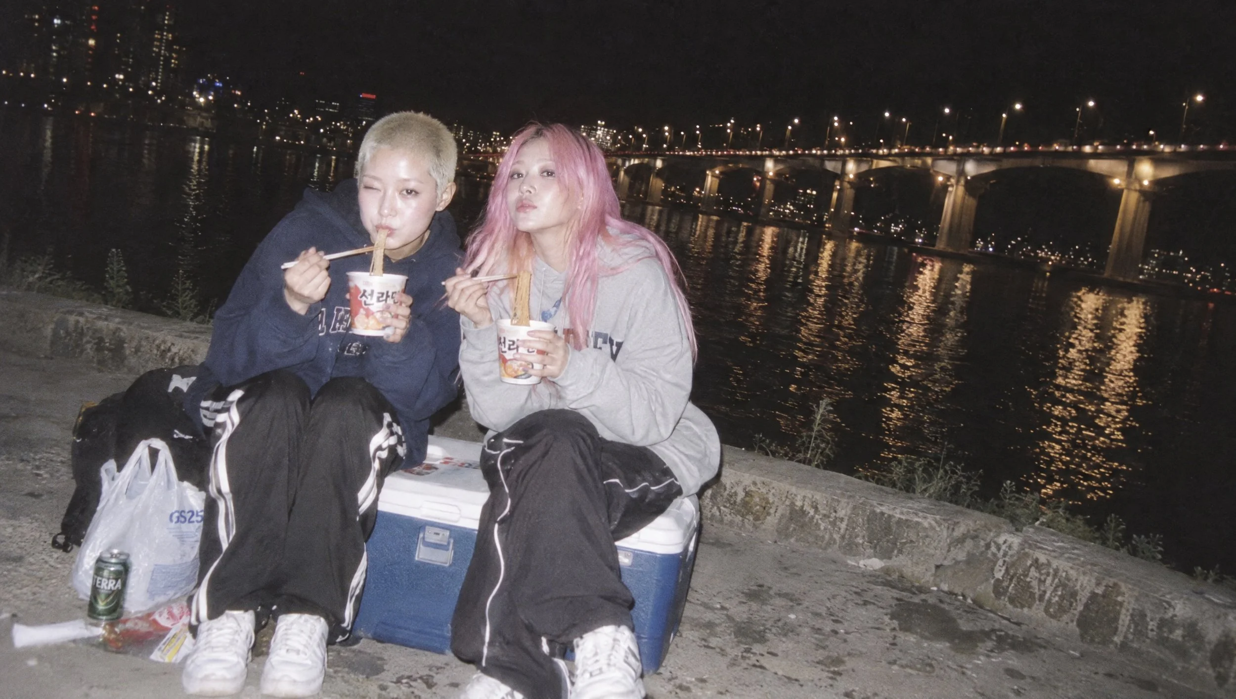 Two friends eating cup ramen by a river at night.