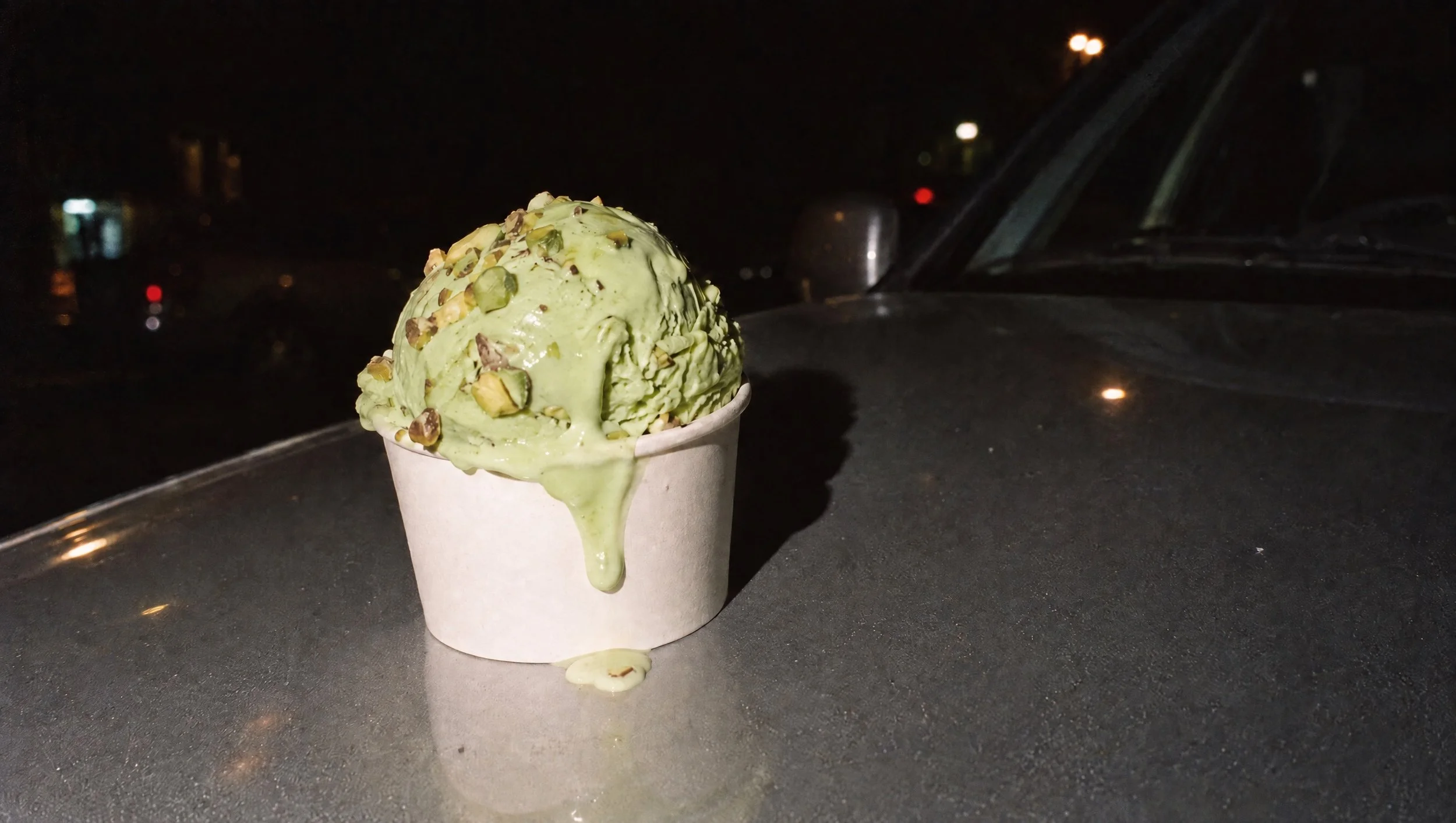 A melting pistachio ice cream cup on a car roof at night, shot with direct flash.