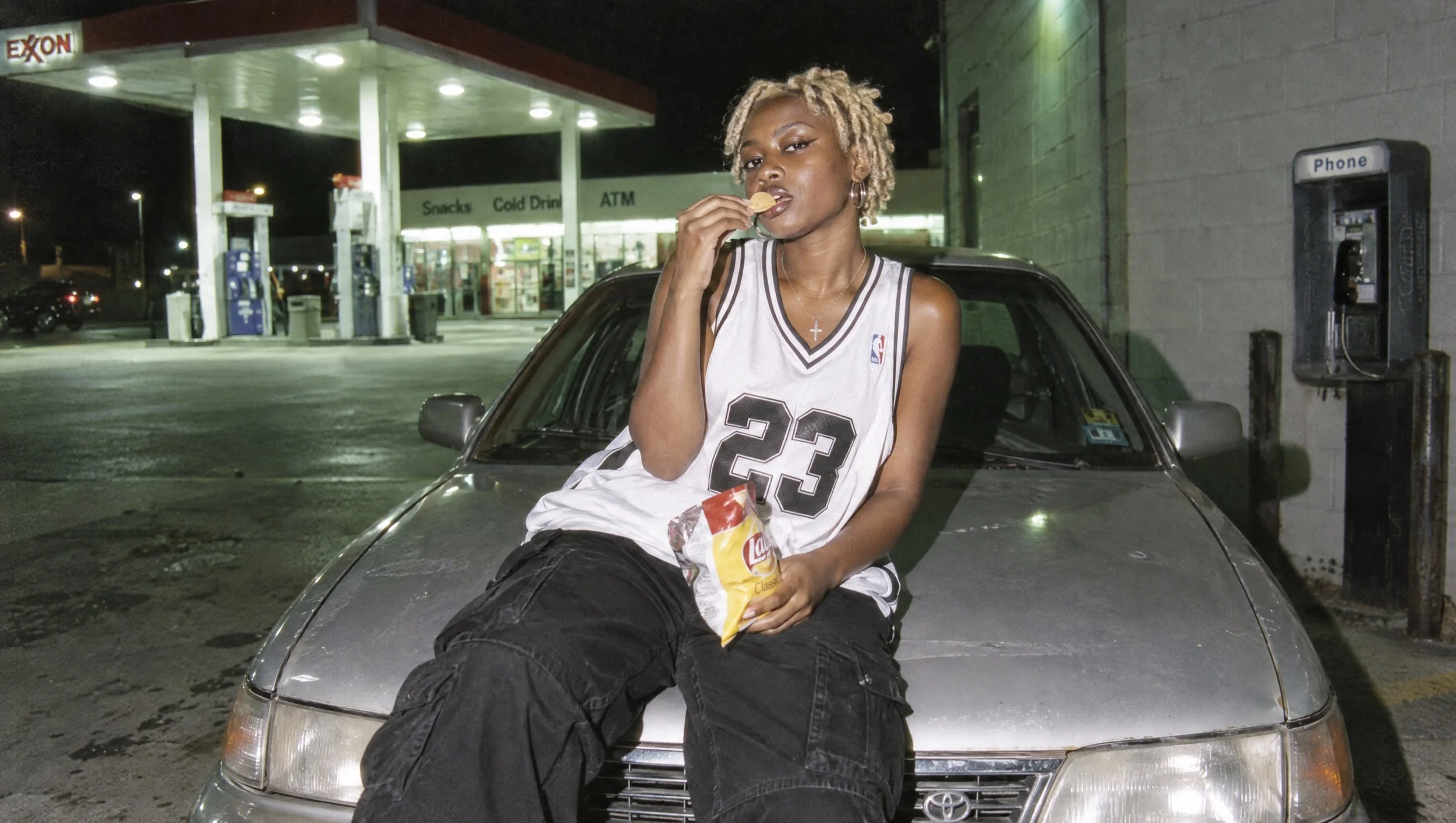A young man sitting on a car hood at a gas station at night.