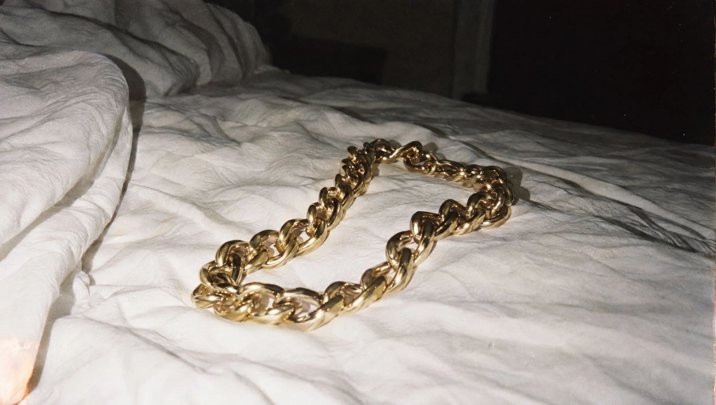 A heavy gold chain necklace on rumpled white sheets, shot with direct flash.