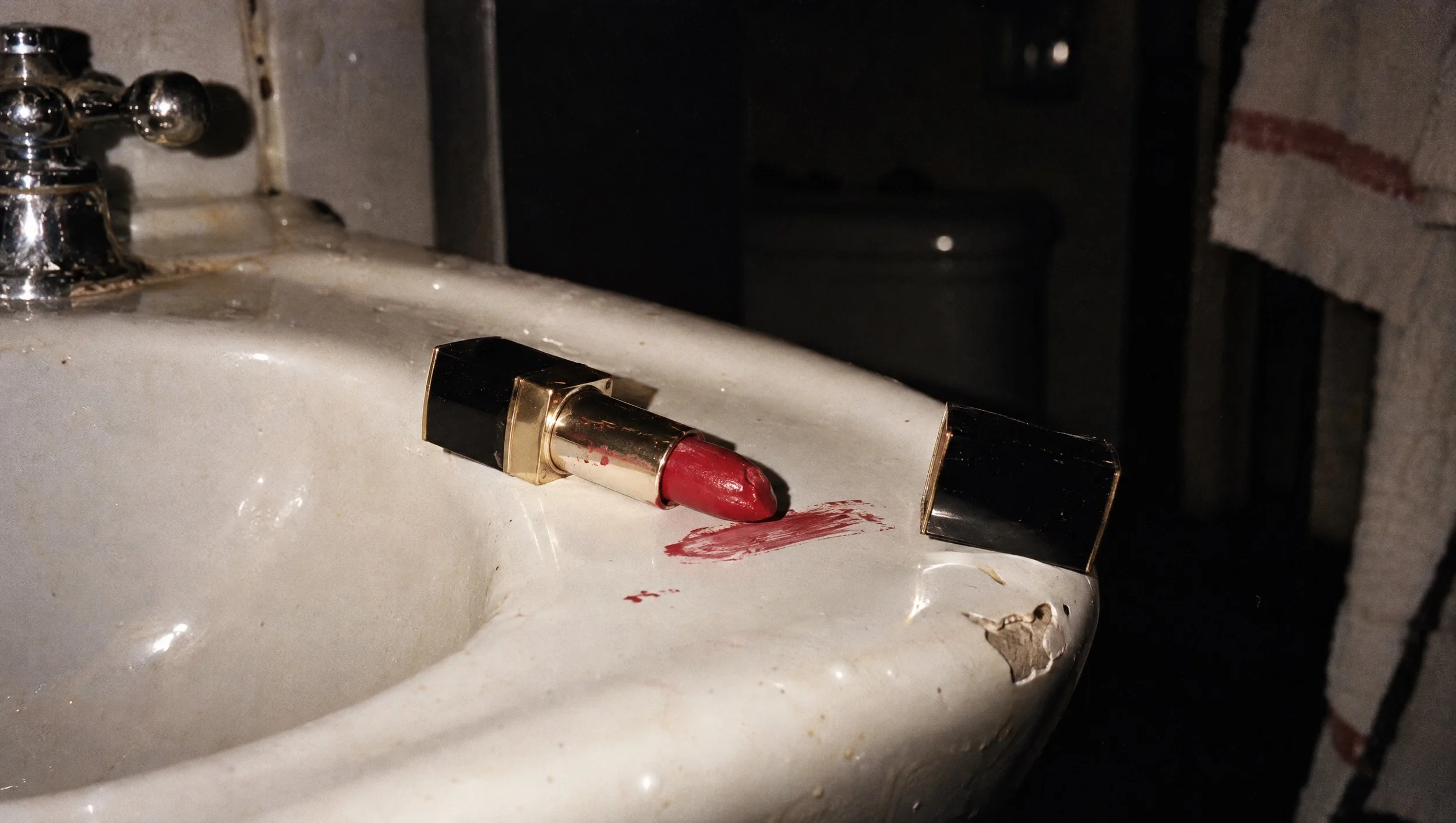 A red lipstick smeared on the edge of a worn bathroom sink, shot with direct flash.