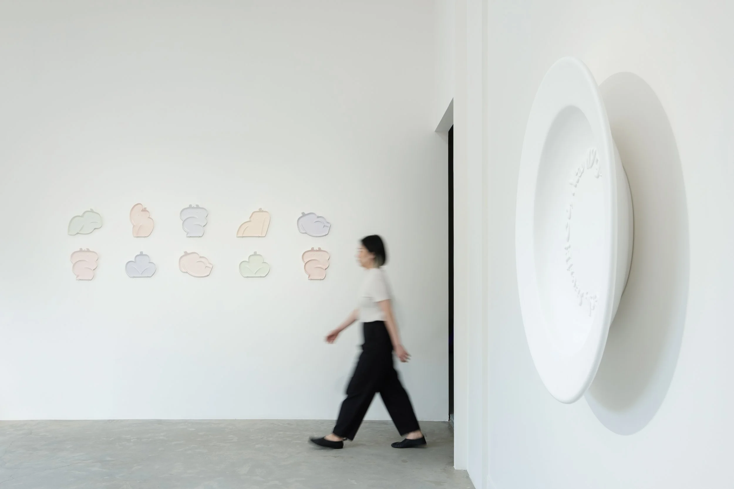 A woman in black pants and a white shirt walking past minimalist art on a white gallery wall, with a large white oval-shaped sculpture on the right.
