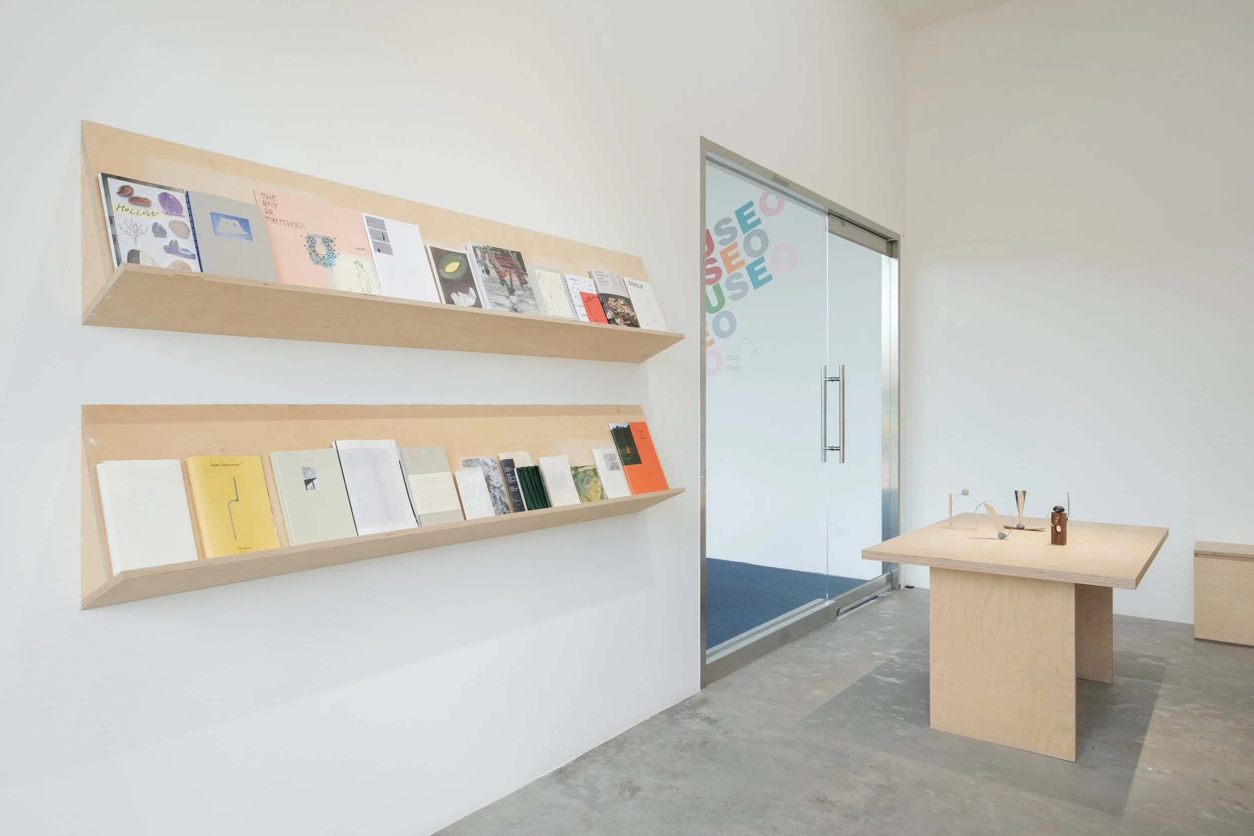 Minimalist interior with two wooden shelves holding books on a white wall, a wooden table with modern objects, and a glass door with colorful text.