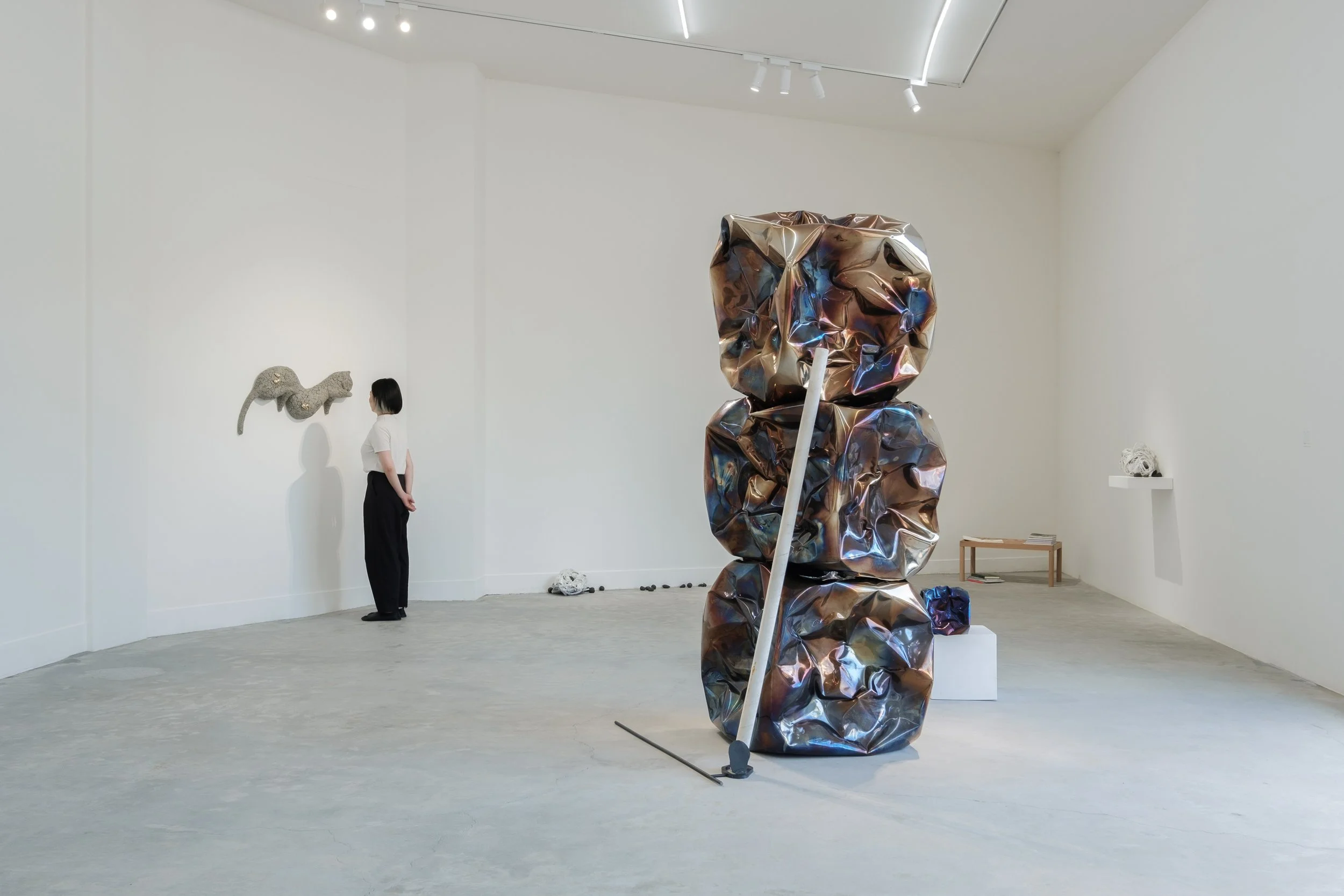An art gallery with a minimalist white interior. In the foreground, a large abstract sculpture of crumpled, metallic-looking material stacked vertically, leaning on a white pedestal. In the background, a woman with short hair, white top, and black pa