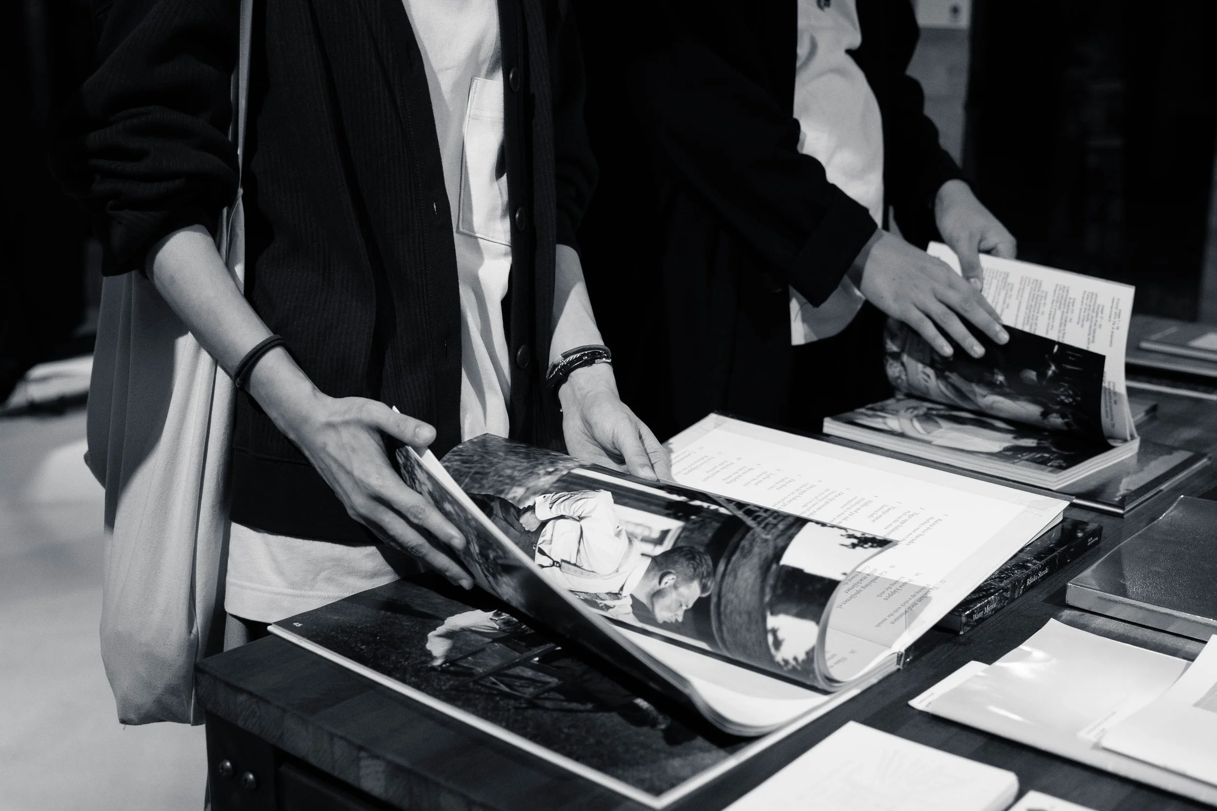 Two people are browsing through magazines and photo books on a table at an indoor event, with only their torsos and hands visible.