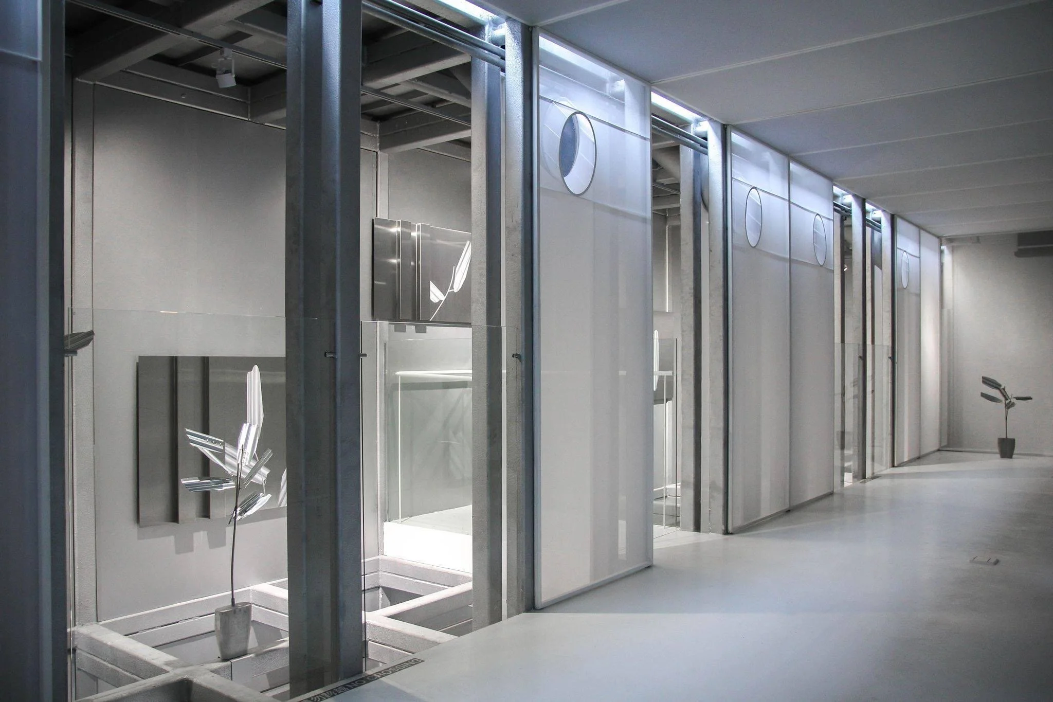 A modern, minimalist indoor space with a row of metallic or glass partitions or cubicles, and a potted plant in the corner.