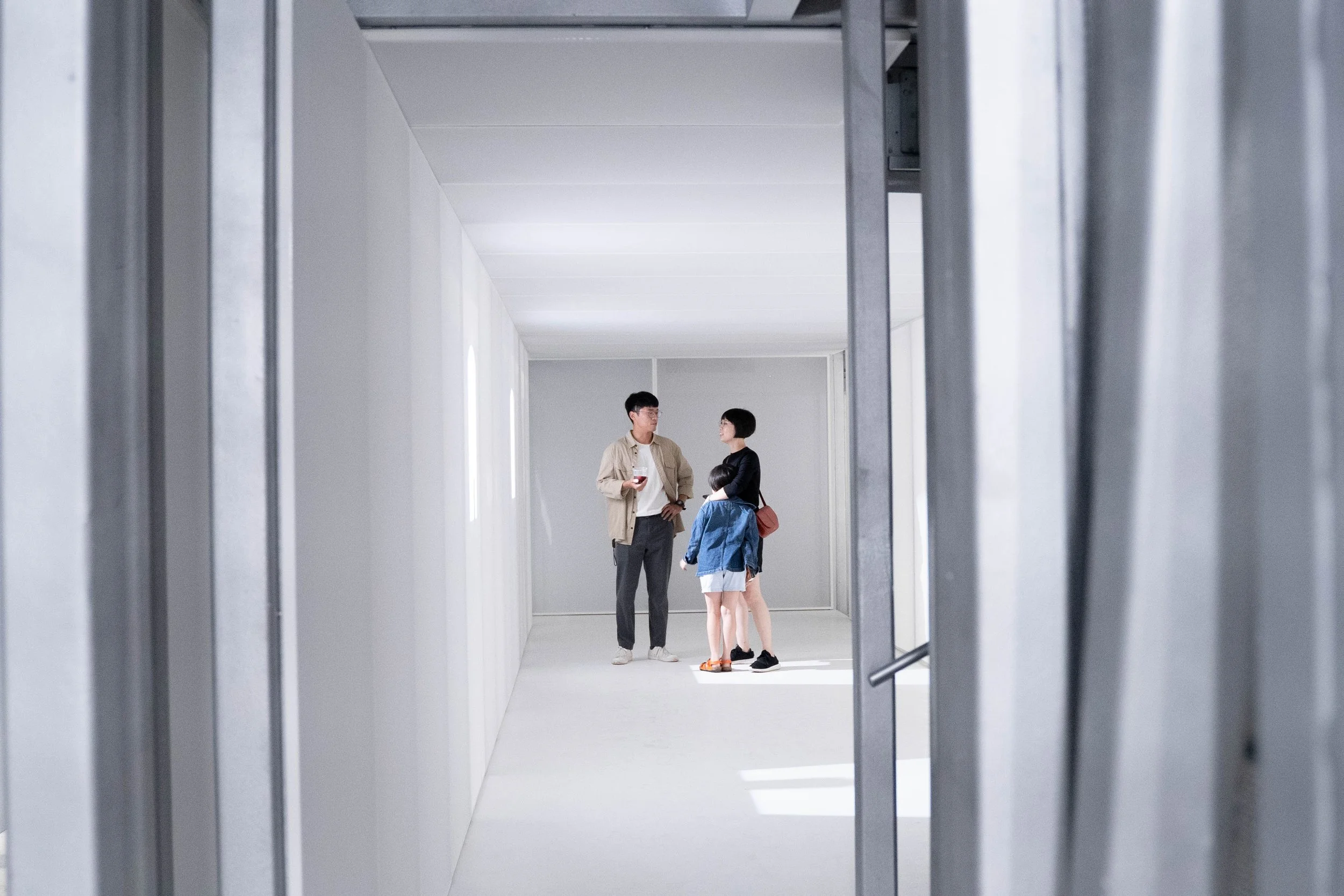 Three people, two adults and a child, standing and talking inside a bright, minimalist, gray-walled room seen through an open metallic door frame.
