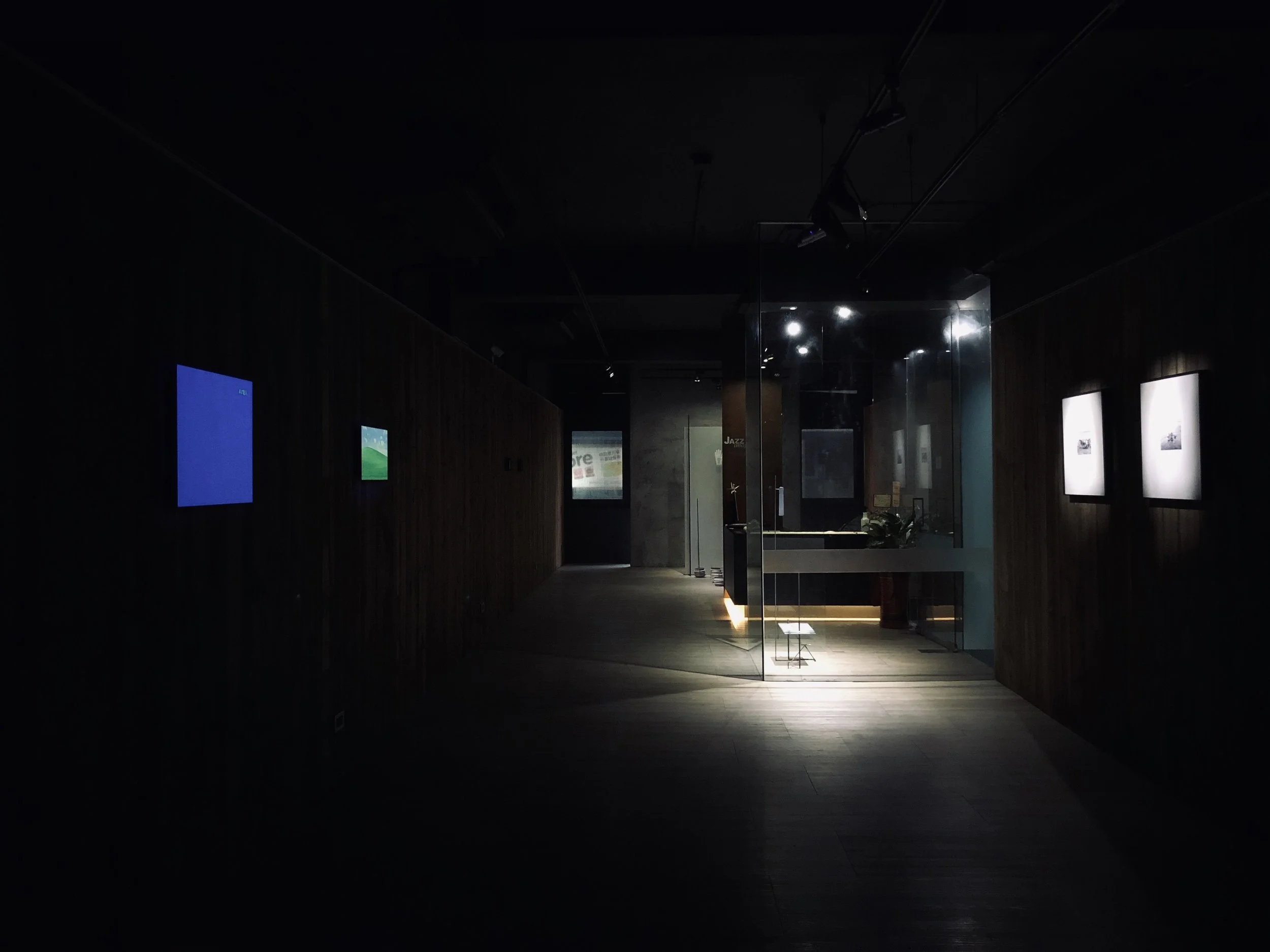 Dimly lit art gallery with digital screens on dark wooden walls, illuminated display area with the centerpiece behind glass, and framed artworks on the right wall.