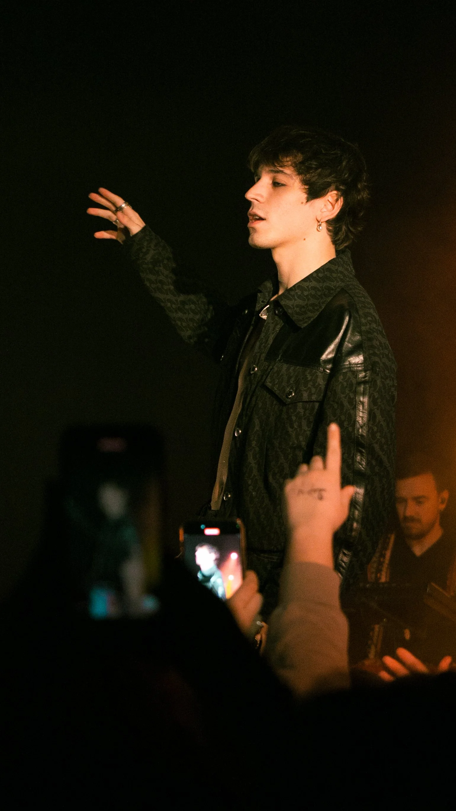 Un giovane uomo con capelli corti e ondulati indossa una giacca nera con dettagli lucidi e sta parlando o cantando, circondato da persone che lo fotografano o registrano. Concerto Alex Wyse