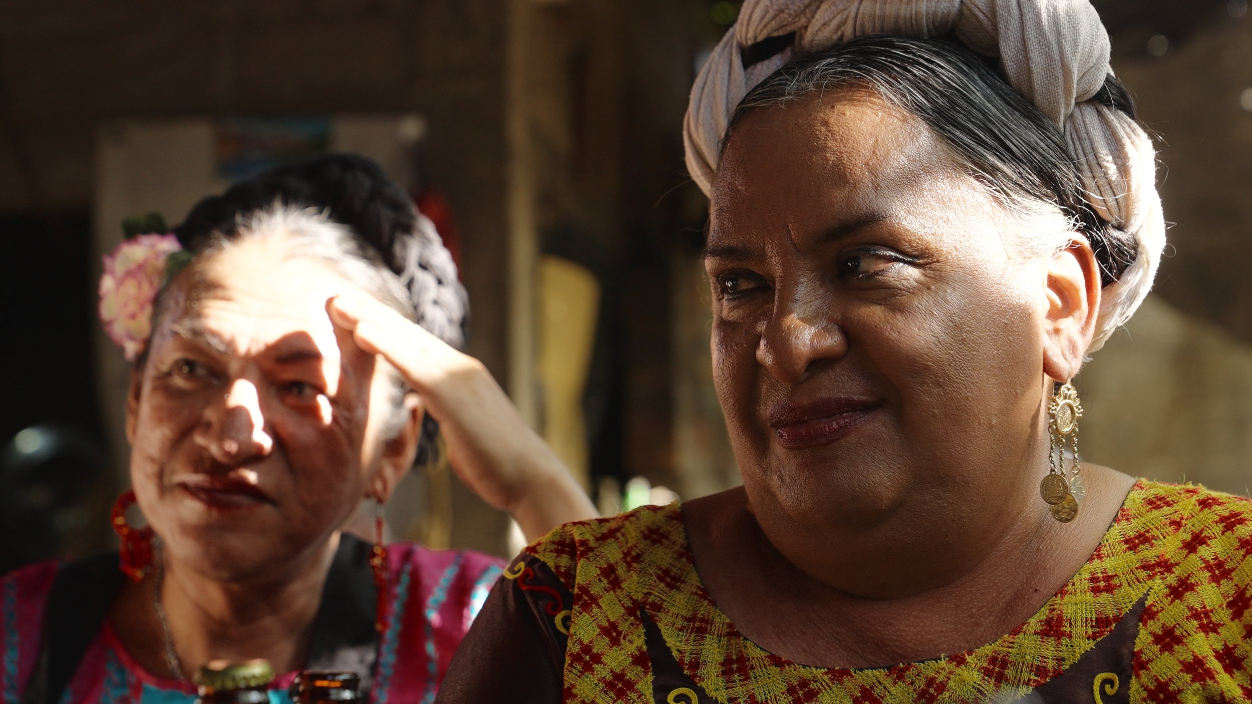 Due donne in abiti tradizionali, una con orecchini pendenti e cappello, l'altra con acconciatura decorata con fiori, in un'atmosfera naturale. Messico Muxe