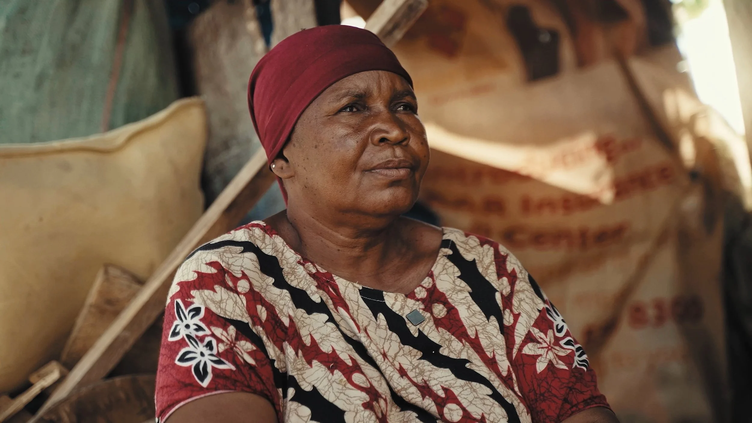 Una donna anziana con pelle scura, indossa un turbante rosso e una maglietta colorata con motivi floreali, seduta interiormente in un ambiente con tessuti e materiali di recupero sullo sfondo, Tanzania.