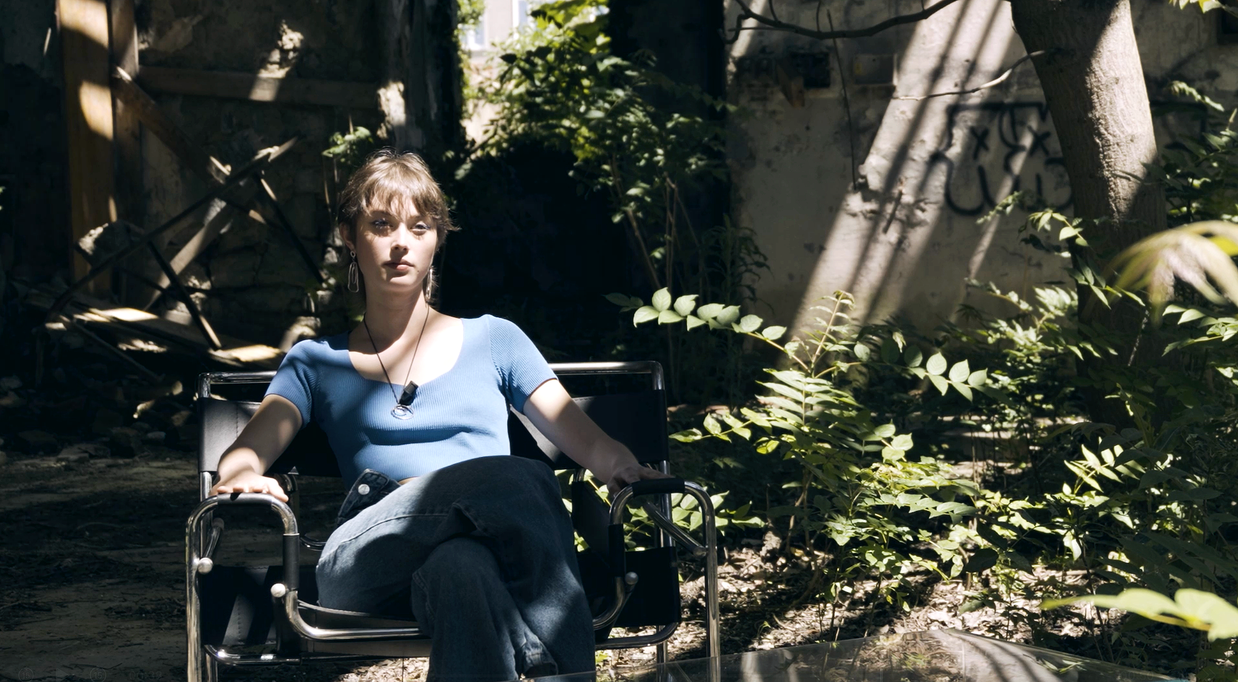 Una ragazza con camicetta blu seduta su una sedia in un'area all'aperto circondata dalla vegetazione, con illuminazione solare che crea ombre e luci