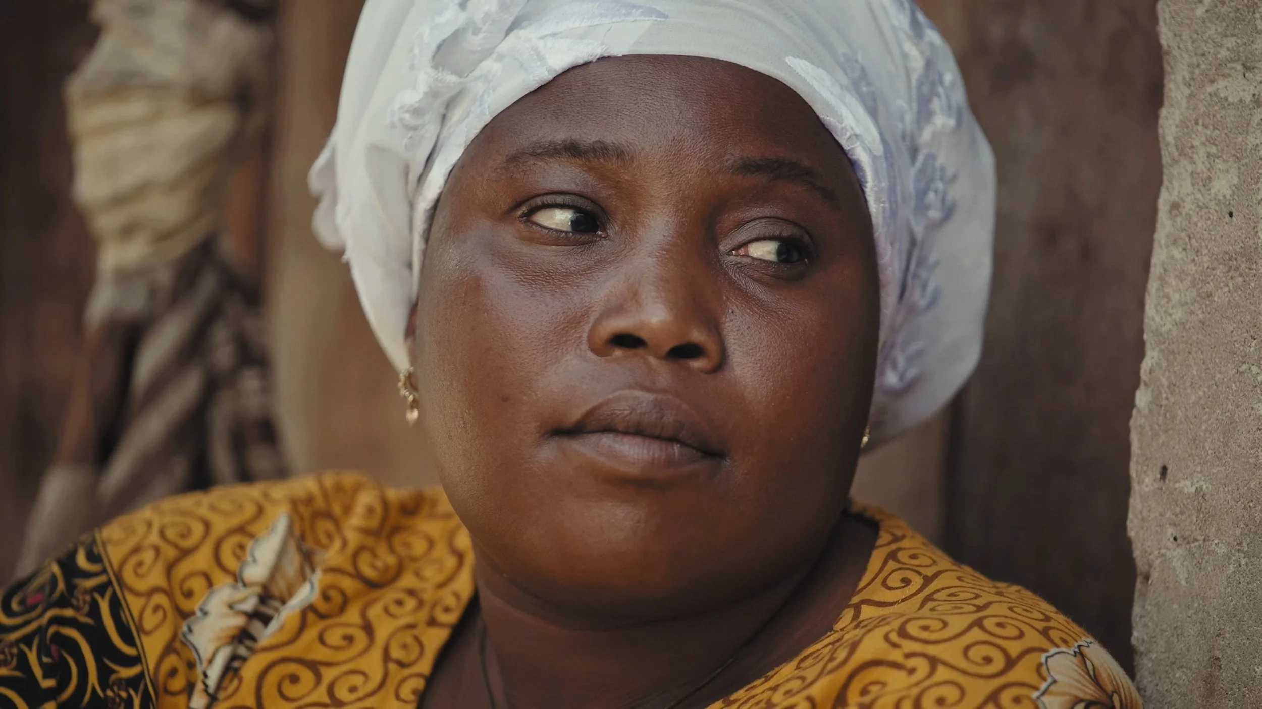 Ragazza africana che indossa un turbante bianco con motivi, rossa e gialla, vestita con un abito giallo con motivi neri, seduta di fronte a una parete di pietra. Tanzania