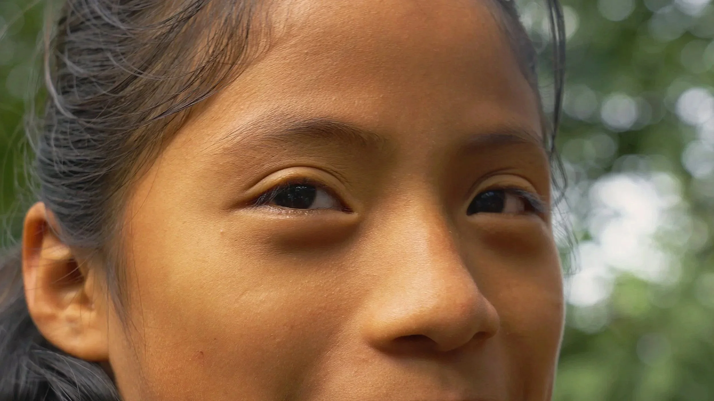 Primo piano del volto di una ragazza asiatica con capelli scuri, occhi a mandorla e pelle luminosa, sfondo sfocato di alberi verdi, Guatemala