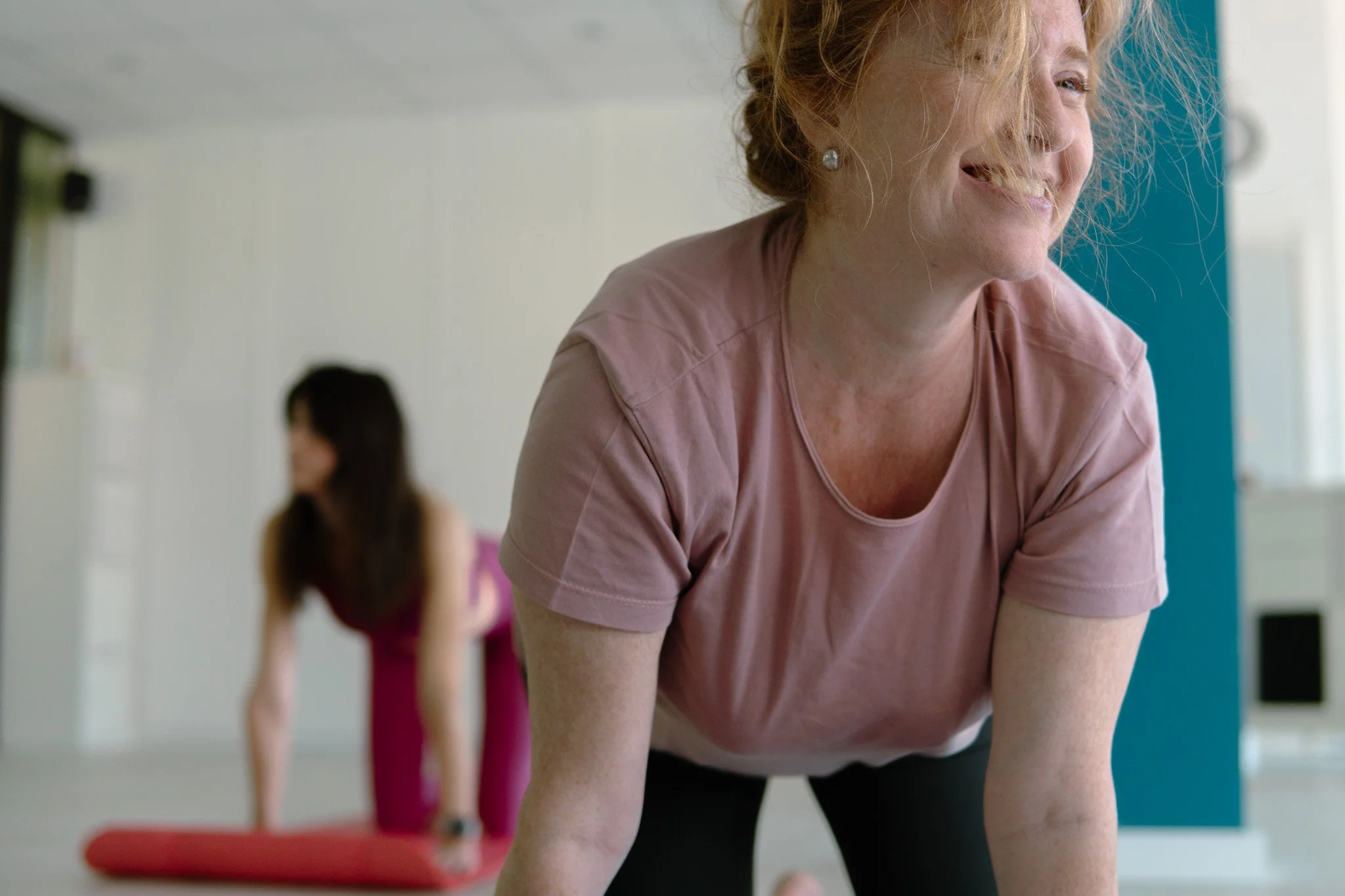 Due donne fanno yoga in una stanza luminosa, una in primo piano sorridente e l'altra in background in posizione di acrobazia. YOGA