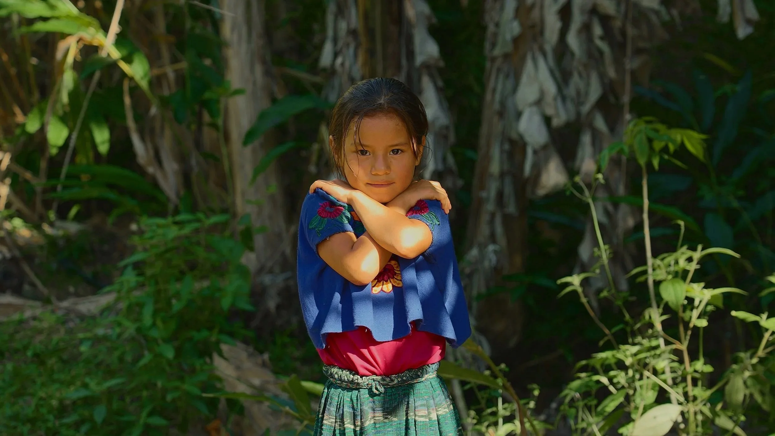 Una bambina con camicia blu con ricami colorati si trova in un ambiente naturale con vegetazione, Comunità Maya in Guatemala