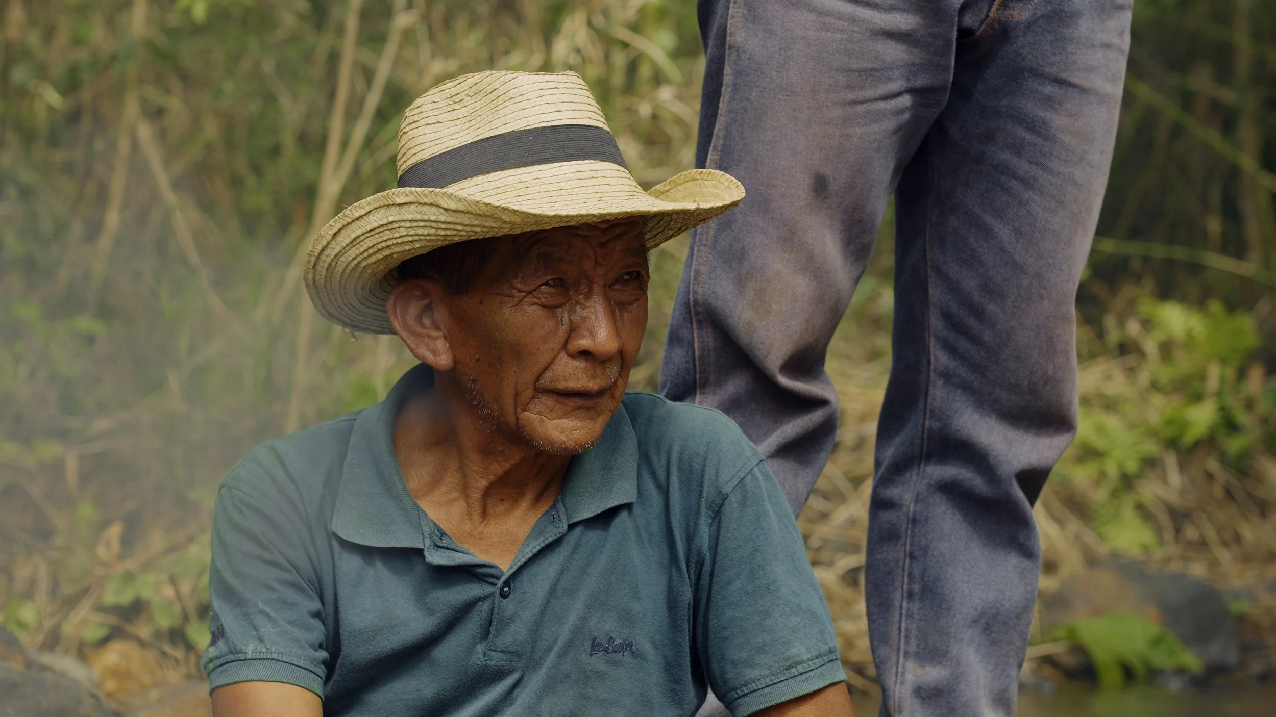 Uomo anziano con cappello di paglia, seduto all'aperto tra vegetazione e fango, rito maya in Guatemala