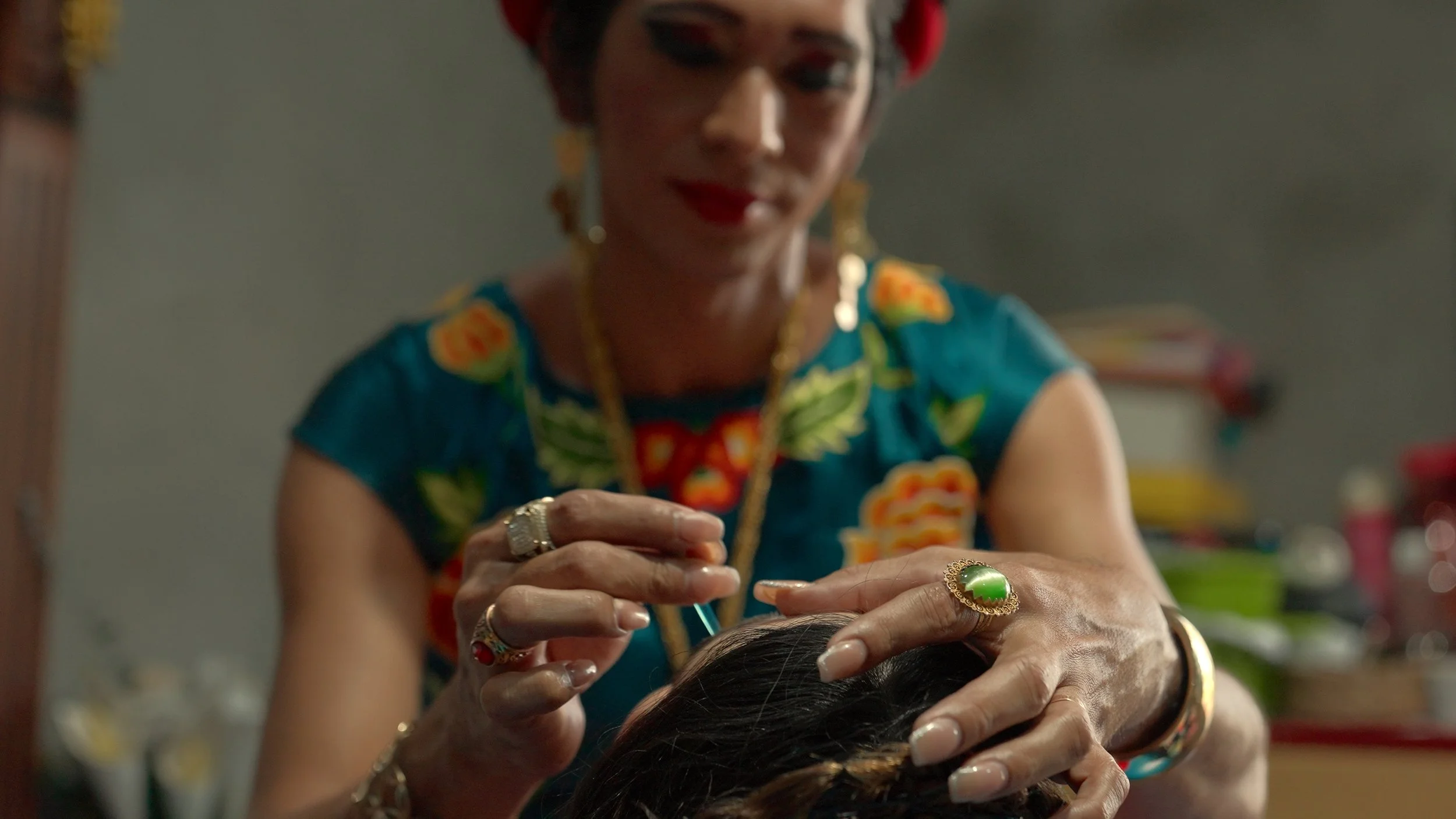 Una donna anziana con abiti colorati, gioielli e capelli corti, applica dell'olio alla testa di una persona che si trova di fronte a lei, in un ambiente domestico. Messico, Muxe
