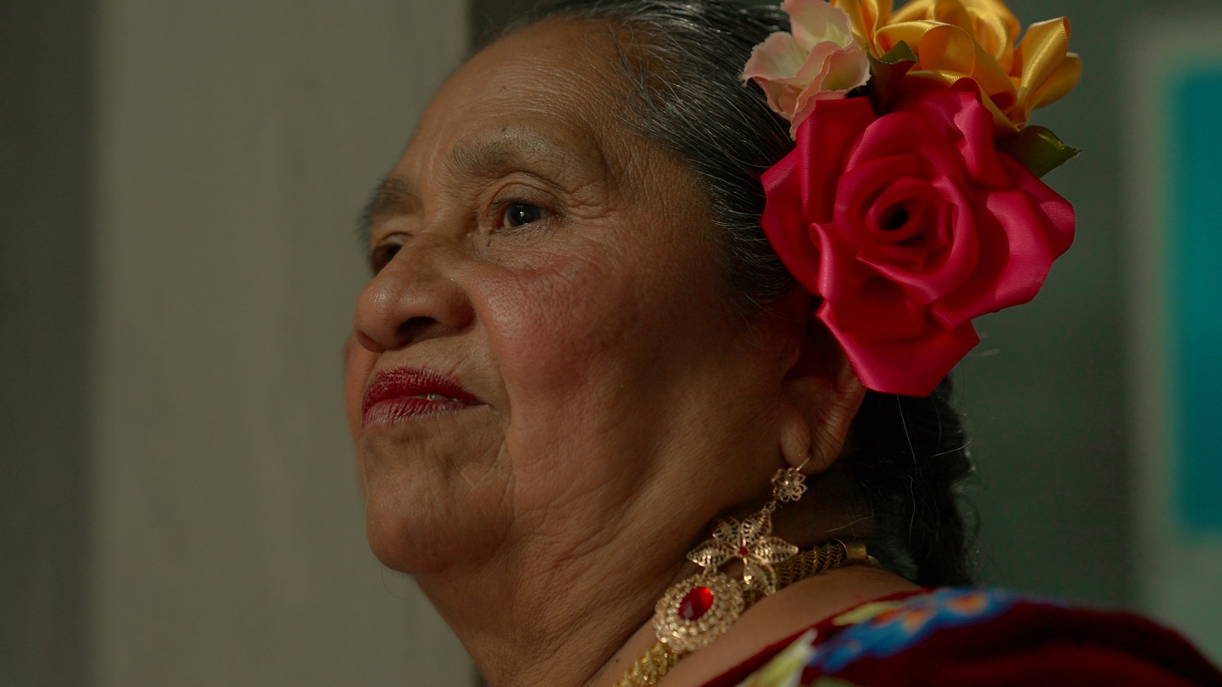Ritratto di una donna anziana con trucco rosso, orecchini d'oro e un fiore rosa e giallo in testa. Messico, Muxe