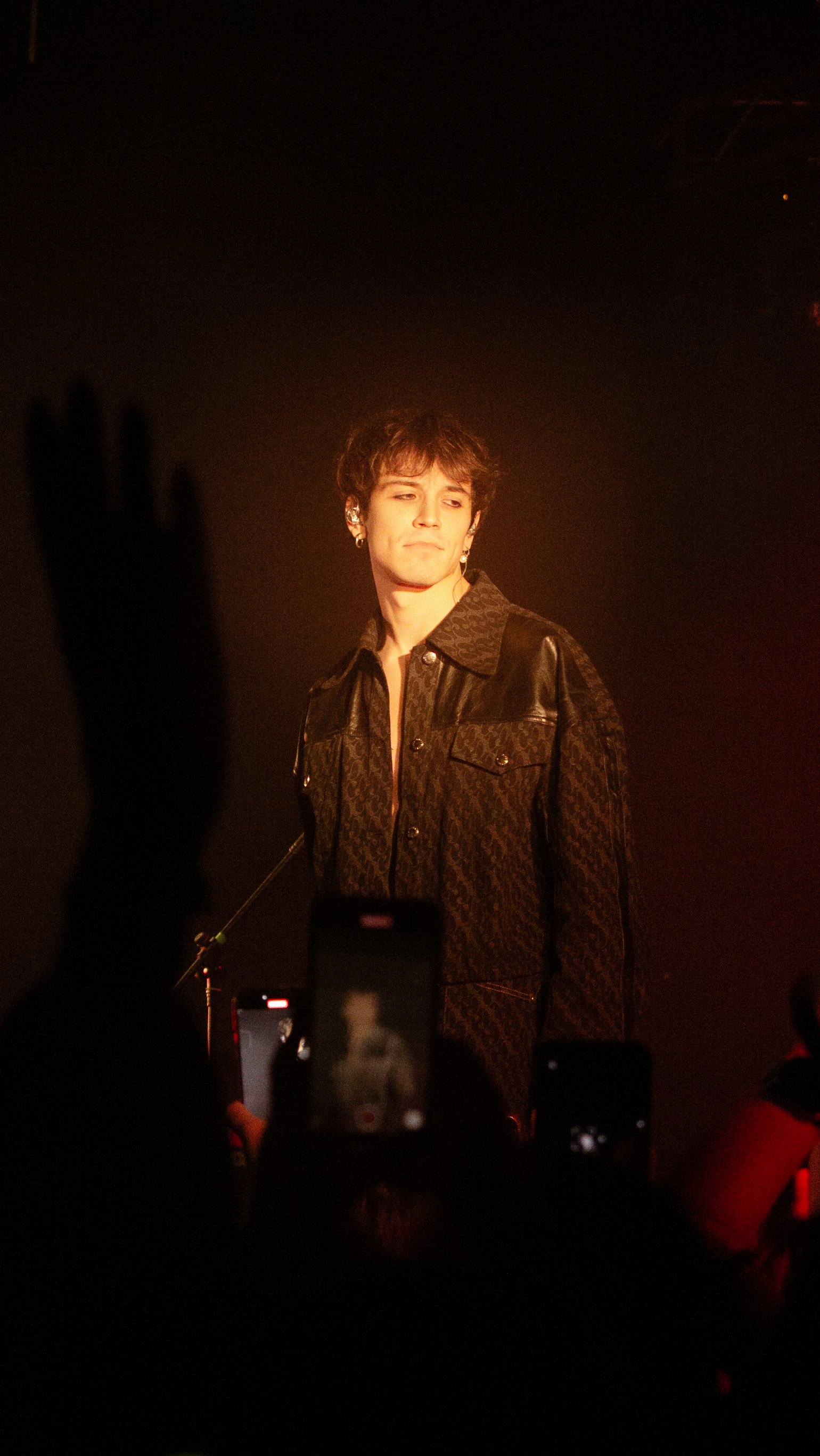 Un giovane artista sul palco durante un concerto, indossa una giacca scura con dettagli in pelle, con luci calde e il pubblico che cattura l'attenzione con i propri cellulari. Concerto Alex Wyse