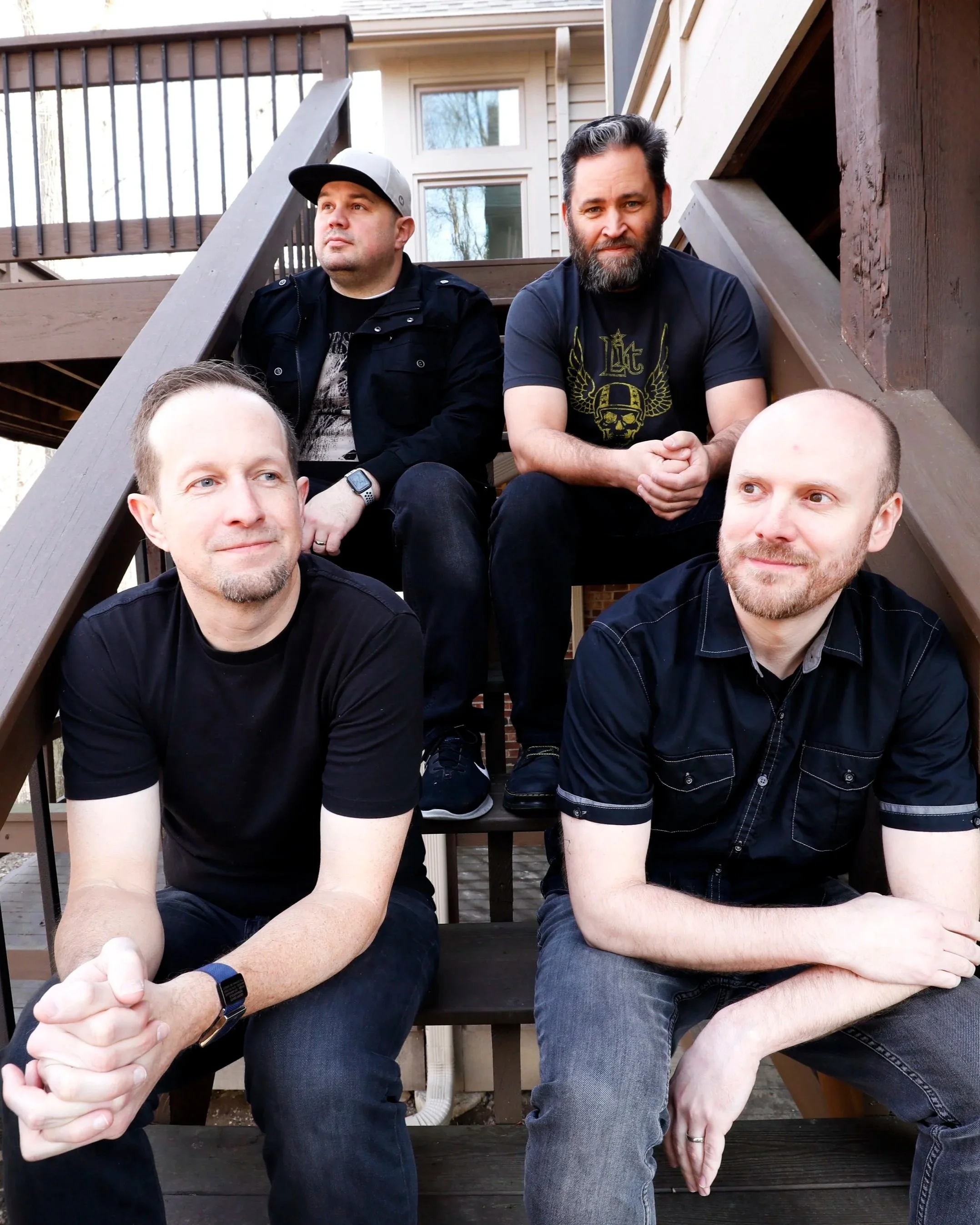 Four men sitting on outdoor wooden stairs, background shows a house with windows and trees.