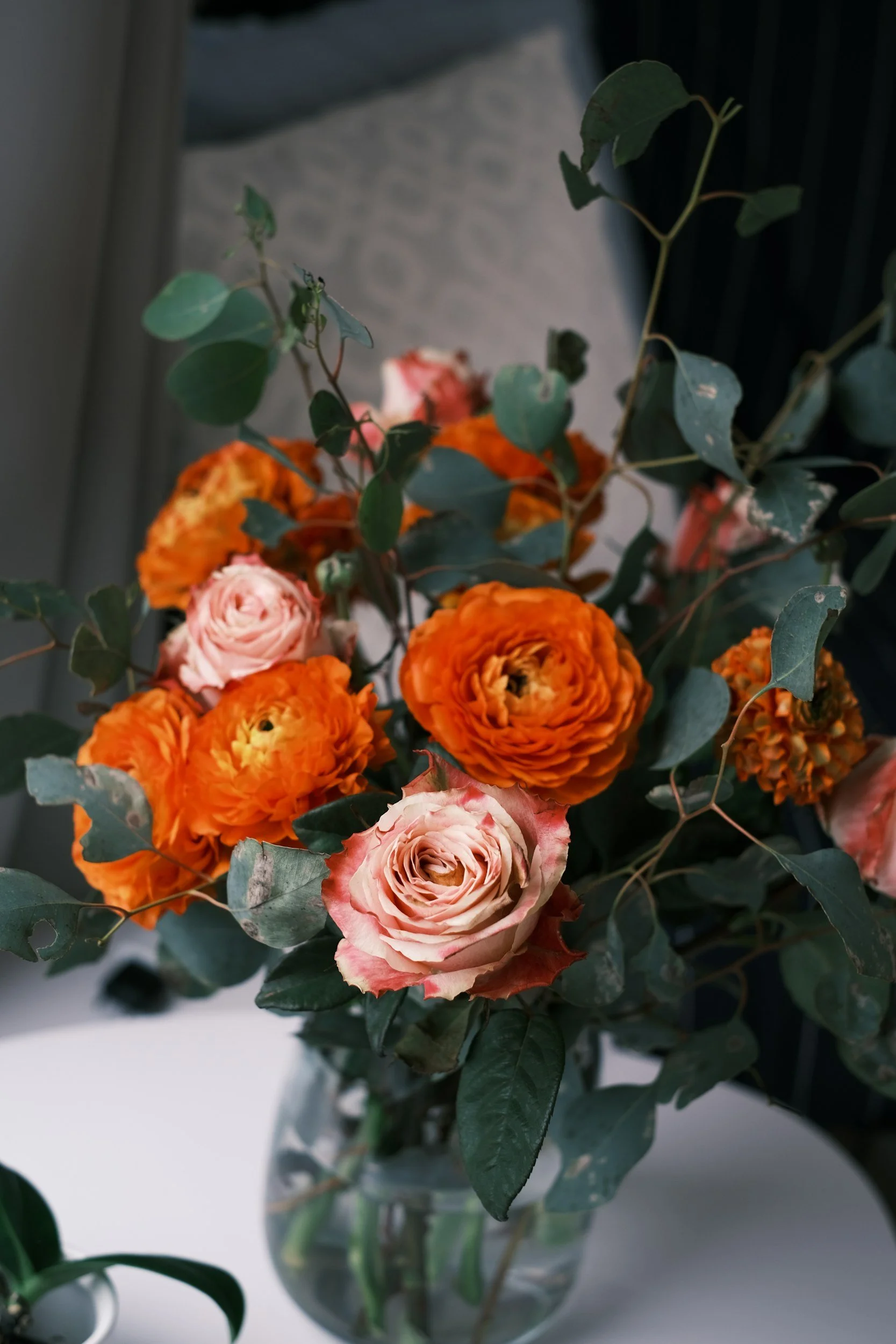 A vase filled with fresh pink and orange flowers, including roses and ranunculus, and green foliage, sitting on a white surface.