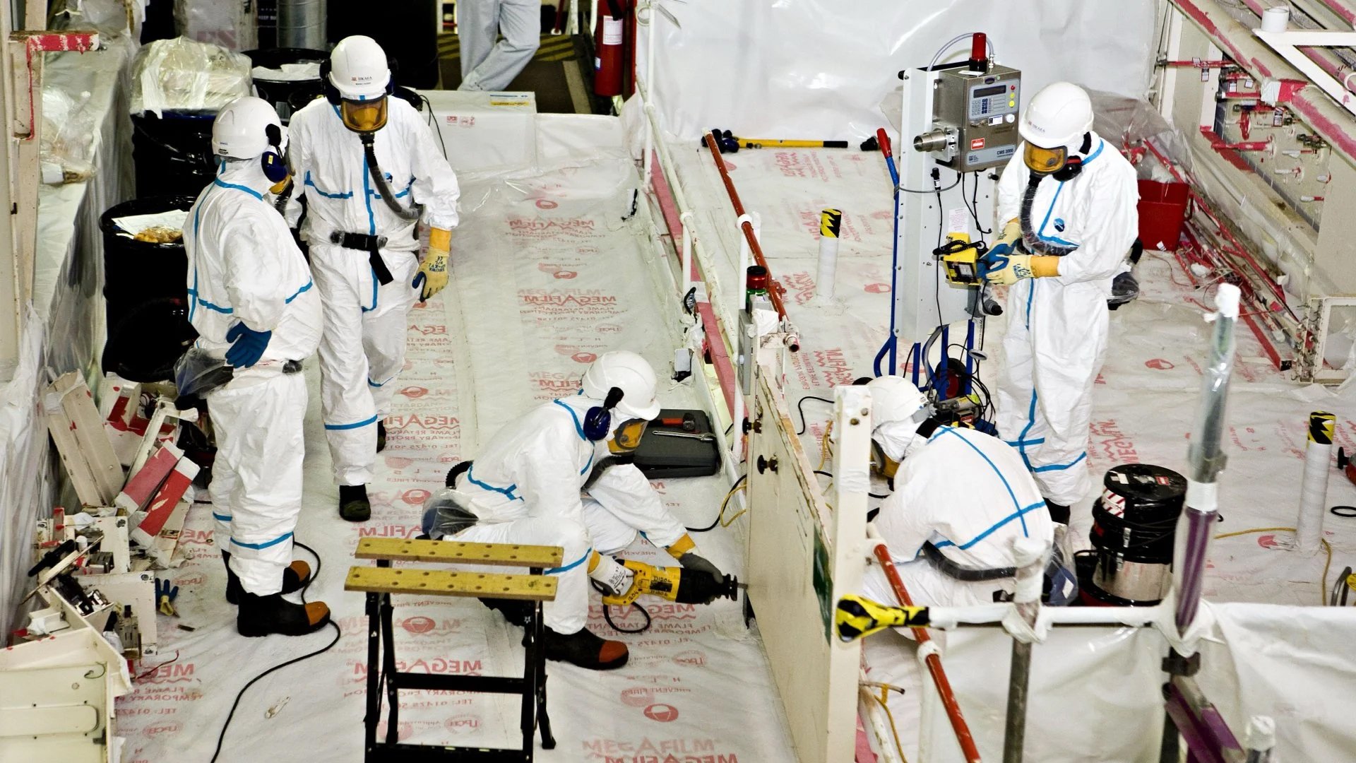 Workers in white protective suits and helmets working on equipment inside a plant room.
