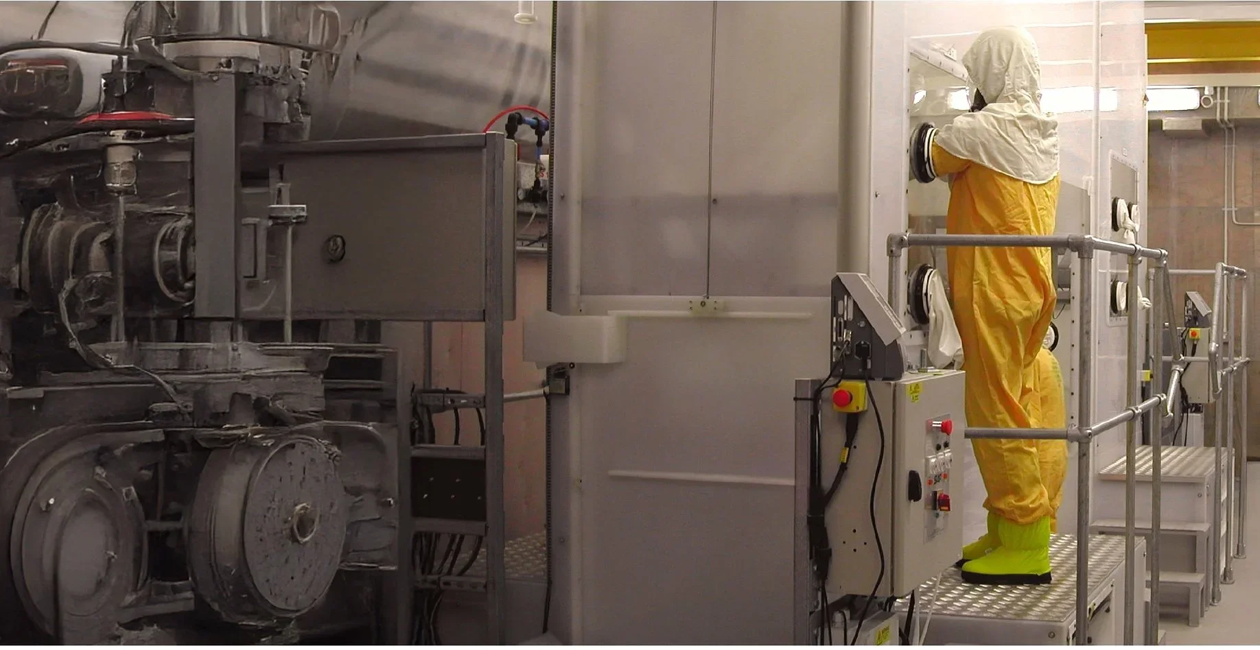 A worker in yellow protective suit and gloves cleaning or inspecting equipment inside industrial facility with machinery and safety railing.