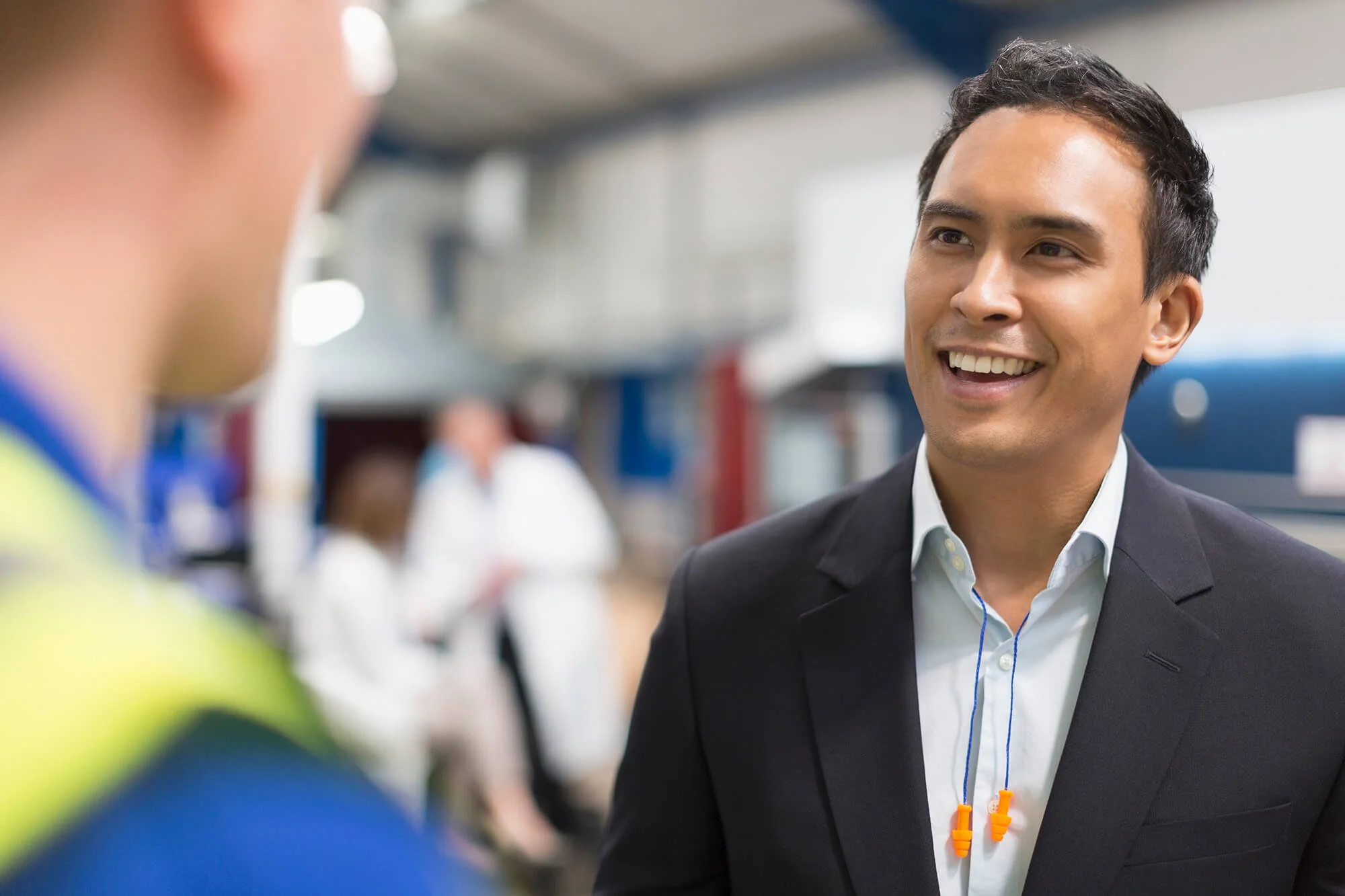 Smiling man in a black suit with earphones talking to a person in a safety vest at an indoor train station.