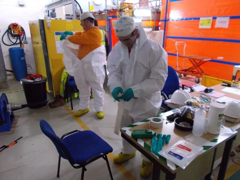 Two people in protective gear working in a laboratory or industrial setting, with one preparing equipment and the other handling supplies.