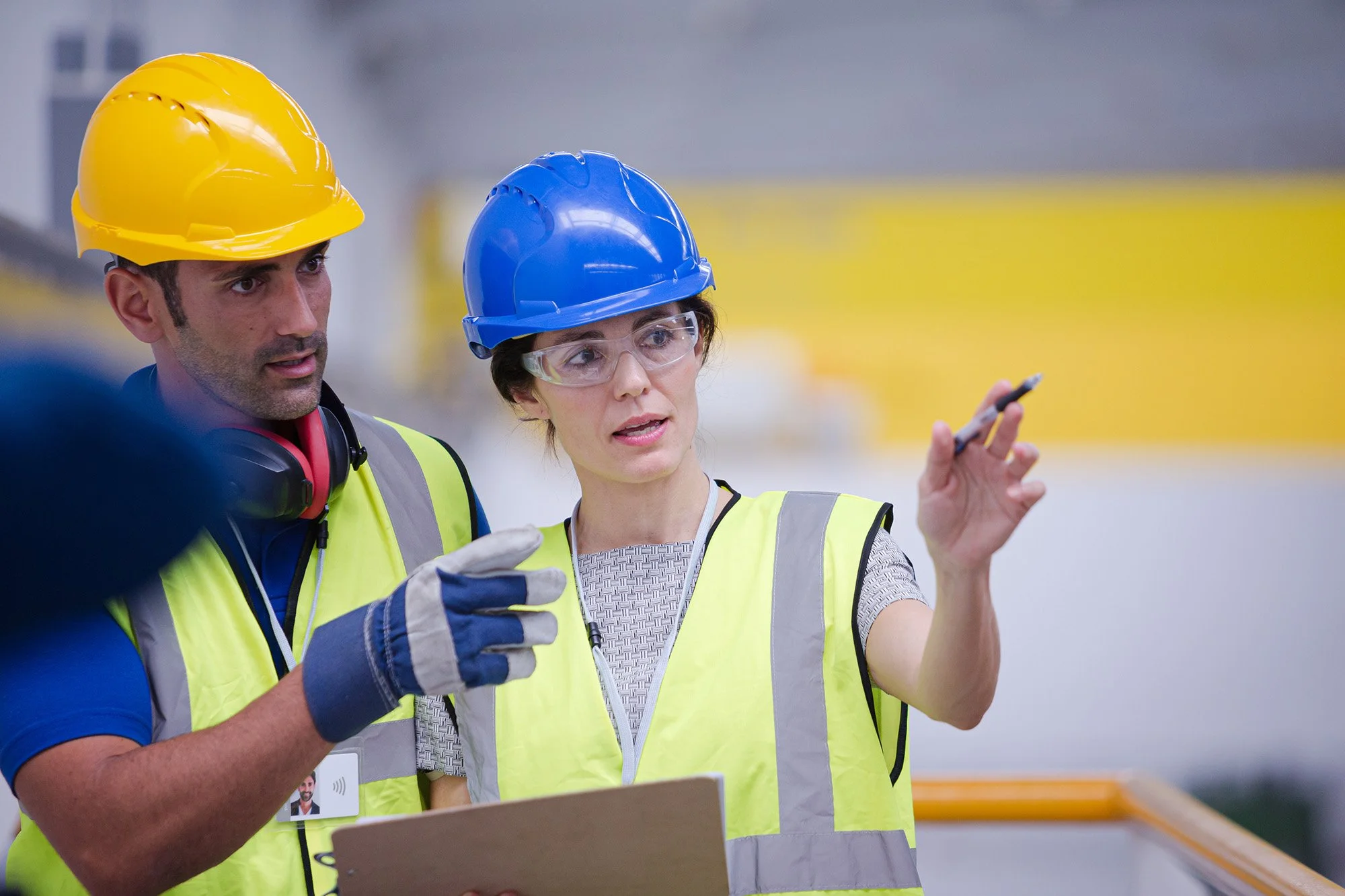 Two plant workers, a man and a woman, wearing safety helmets and high-visibility vests, are discussing on a construction site. The woman is holding a pen and appears to be explaining something, while the man is listening attentively.