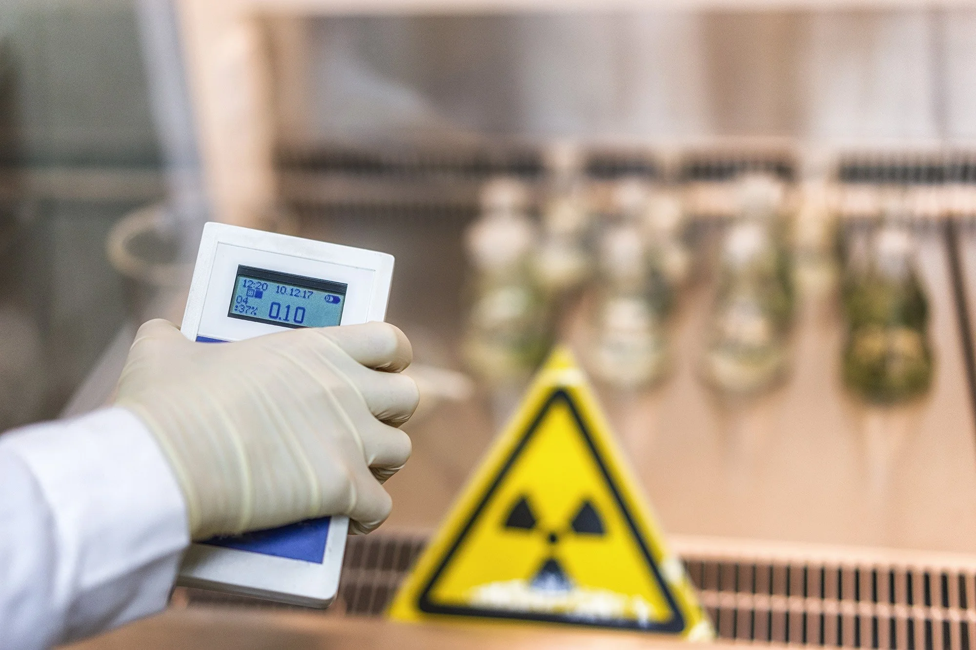 A person wearing gloves holding a digital radiation detector in front of a laboratory fume hood with a nuclear warning sign below it.