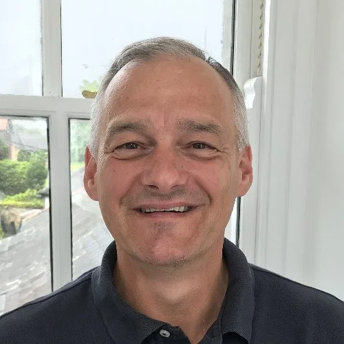 Smiling middle-aged man with short gray hair, wearing a dark collared shirt, standing indoors near a window with a view of trees and rooftops outside.