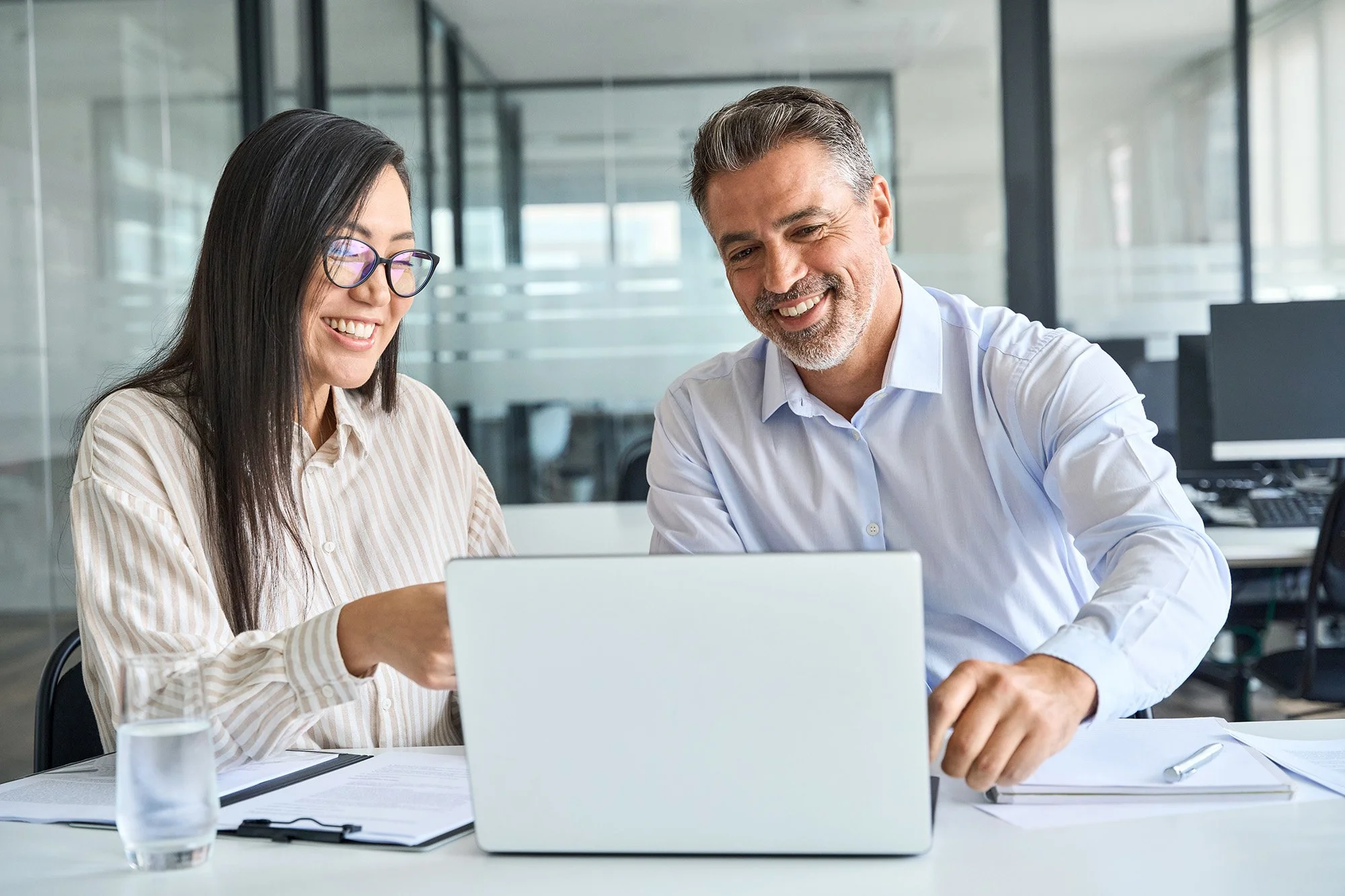 Two professionals, a woman with long black hair and glasses and a man with gray hair and beard, are sitting at a desk in an office, looking at a laptop and smiling.