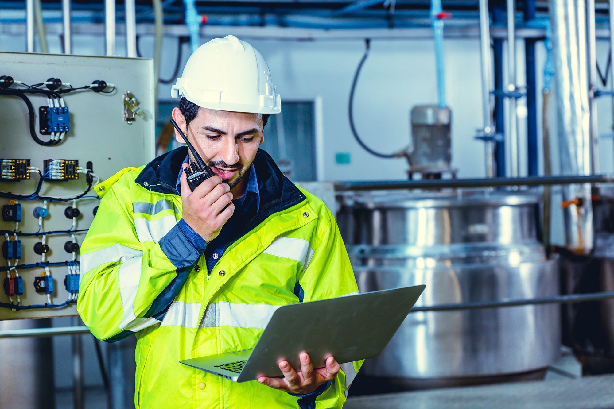 A man wearing a white safety helmet and a bright yellow safety jacket, talking on a walkie-talkie and looking at a laptop in an industrial setting with large metal tanks and control panels in the background.