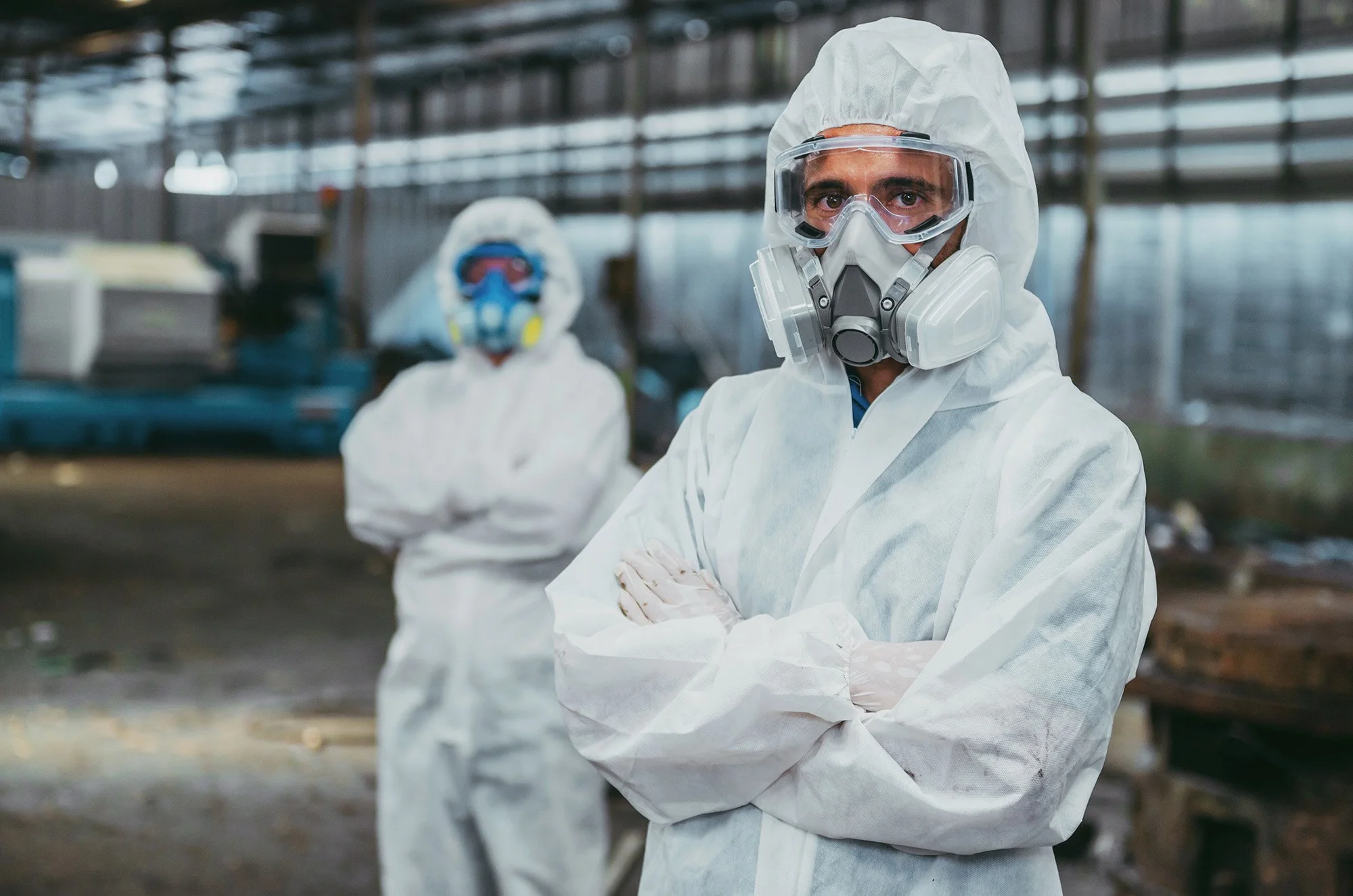 Two people wearing protective suits, masks, and goggles inside a large industrial or research facility.