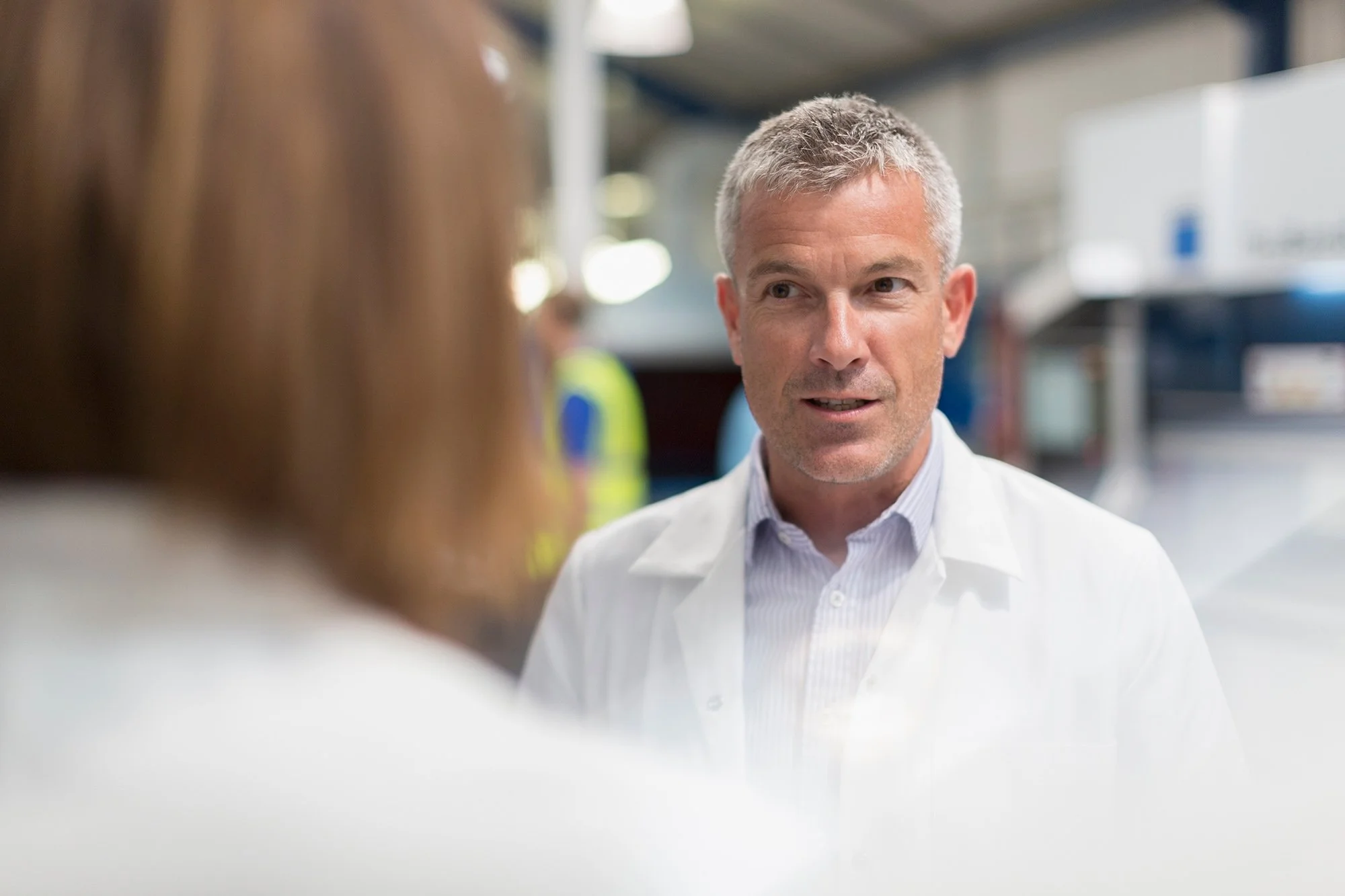 A middle-aged man with short gray hair wearing a white lab coat and a light-colored collared shirt, engaged in a conversation with a woman in an industrial or laboratory setting.