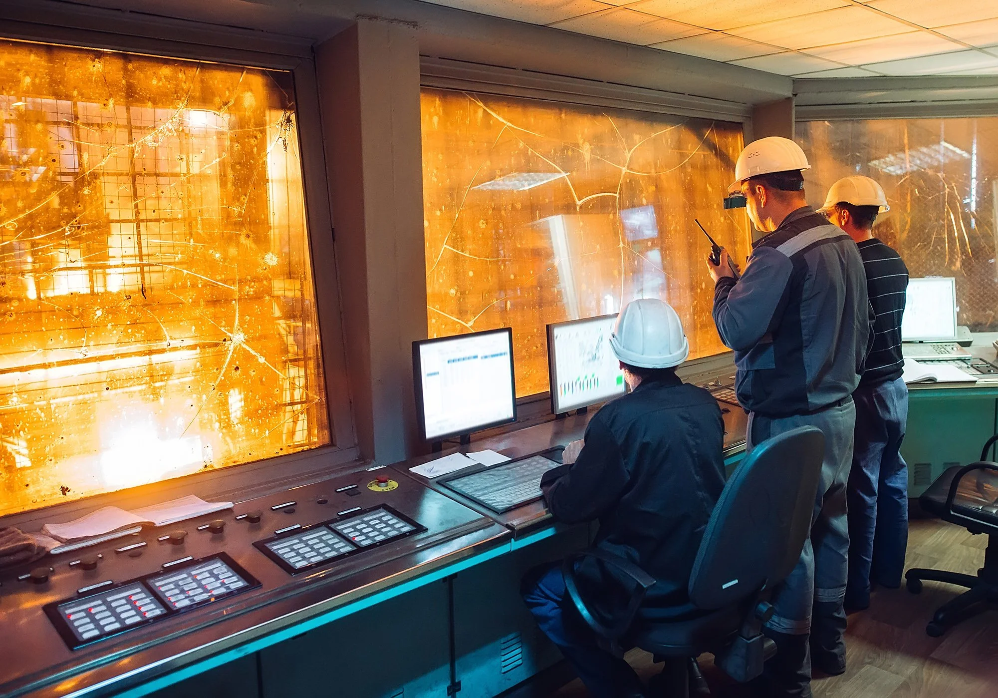Four engineers in hard hats monitor a fire inside a control room with broken, cracked windows.
