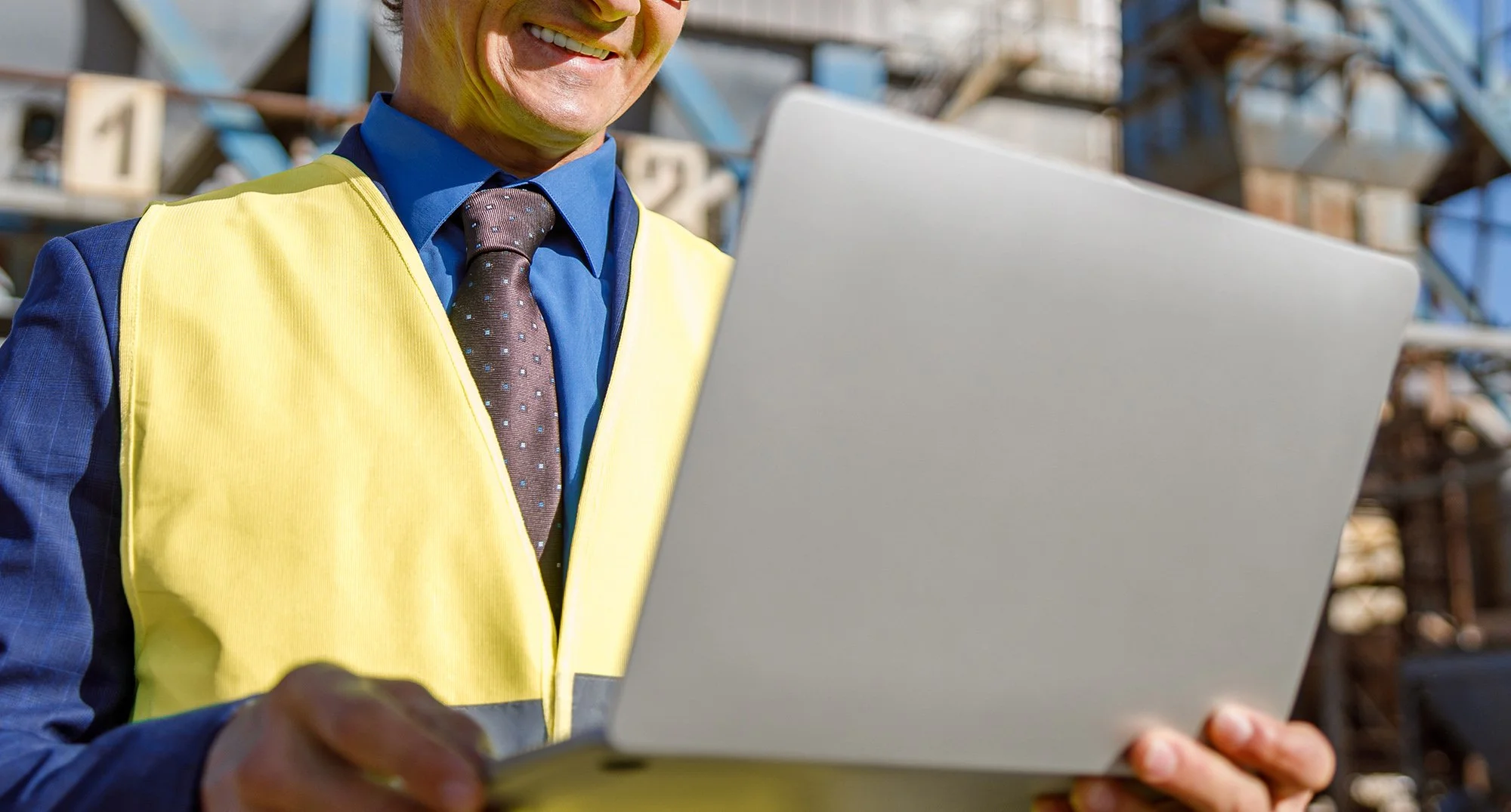A plant worker in a yellow safety vest using a laptop at a plant site.
