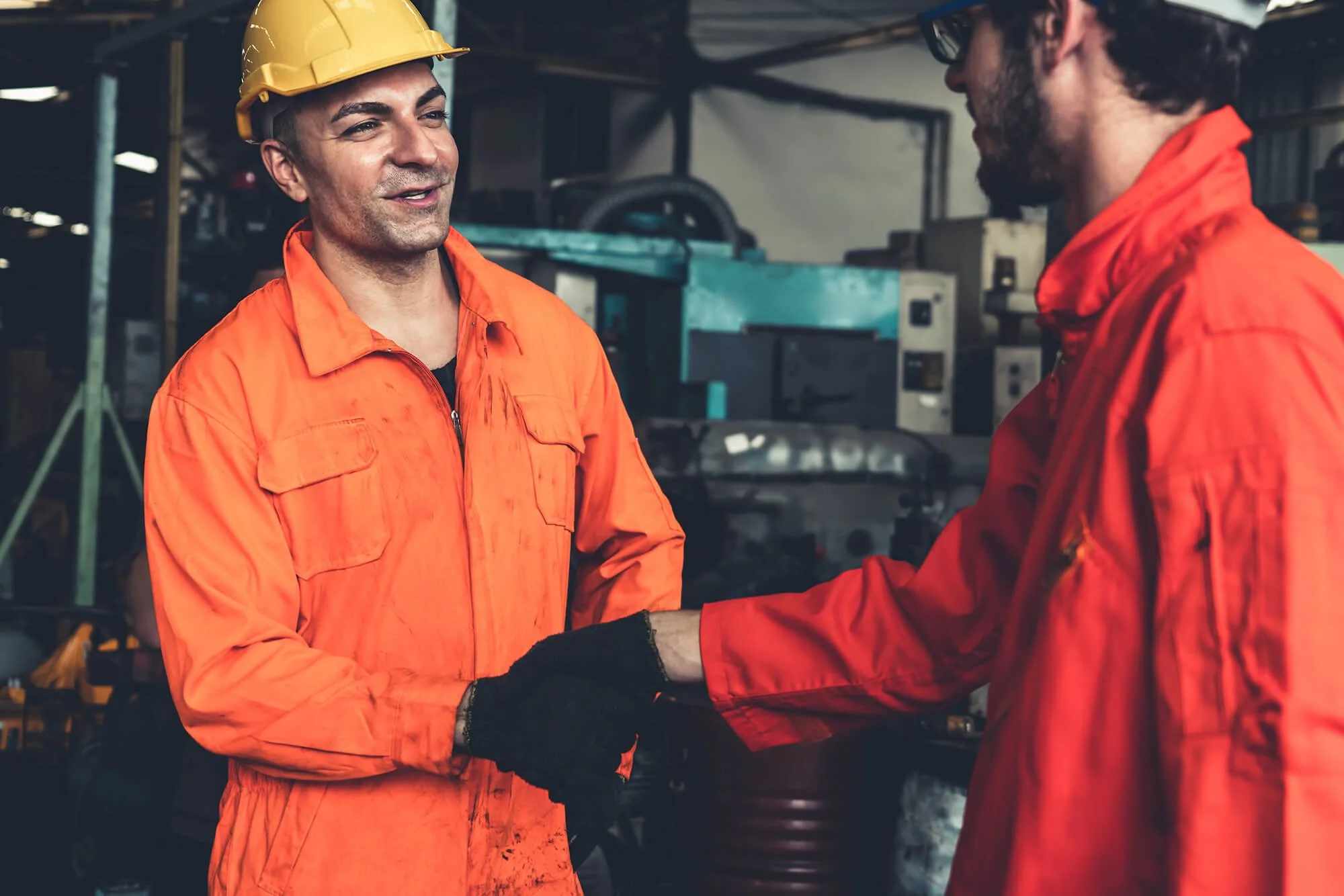 Two workers in orange uniforms shaking hands inside an industrial workshop.