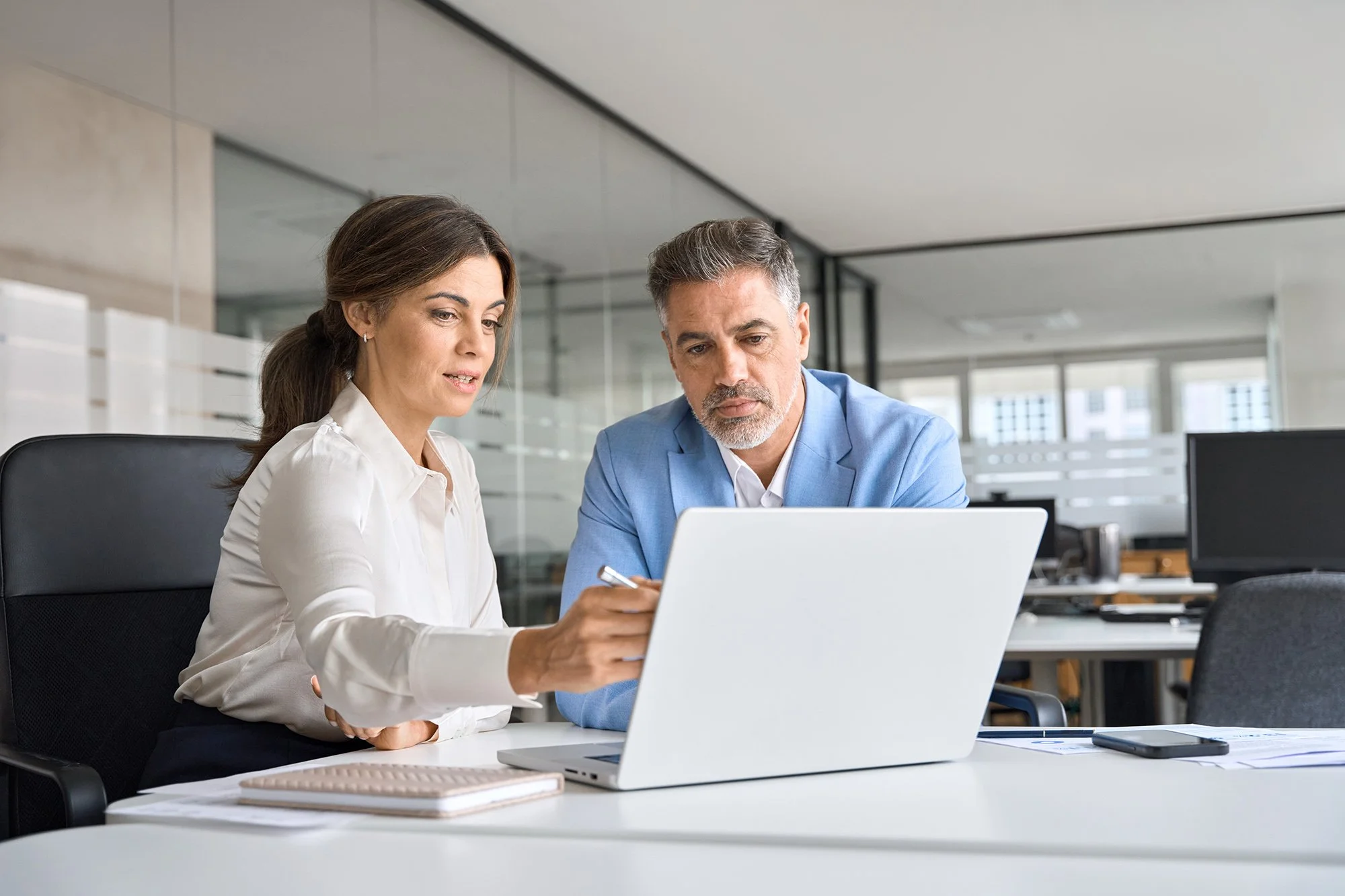 A man and a woman in business attire working together at a table with a laptop in an office.