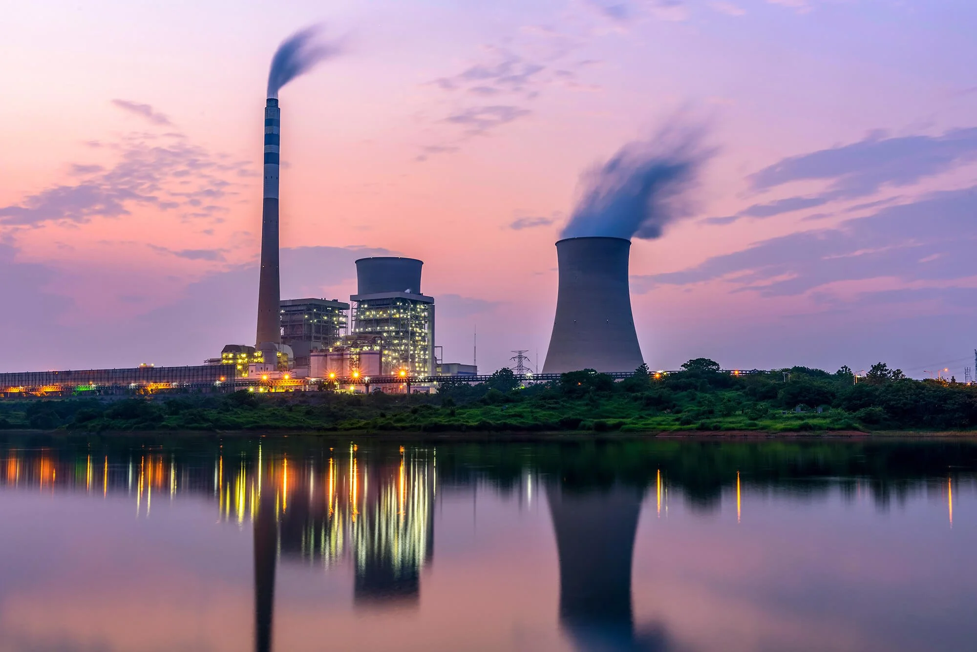 A power plant with cooling towers and a smokestack emitting smoke, reflected on a body of water during sunset or sunrise.